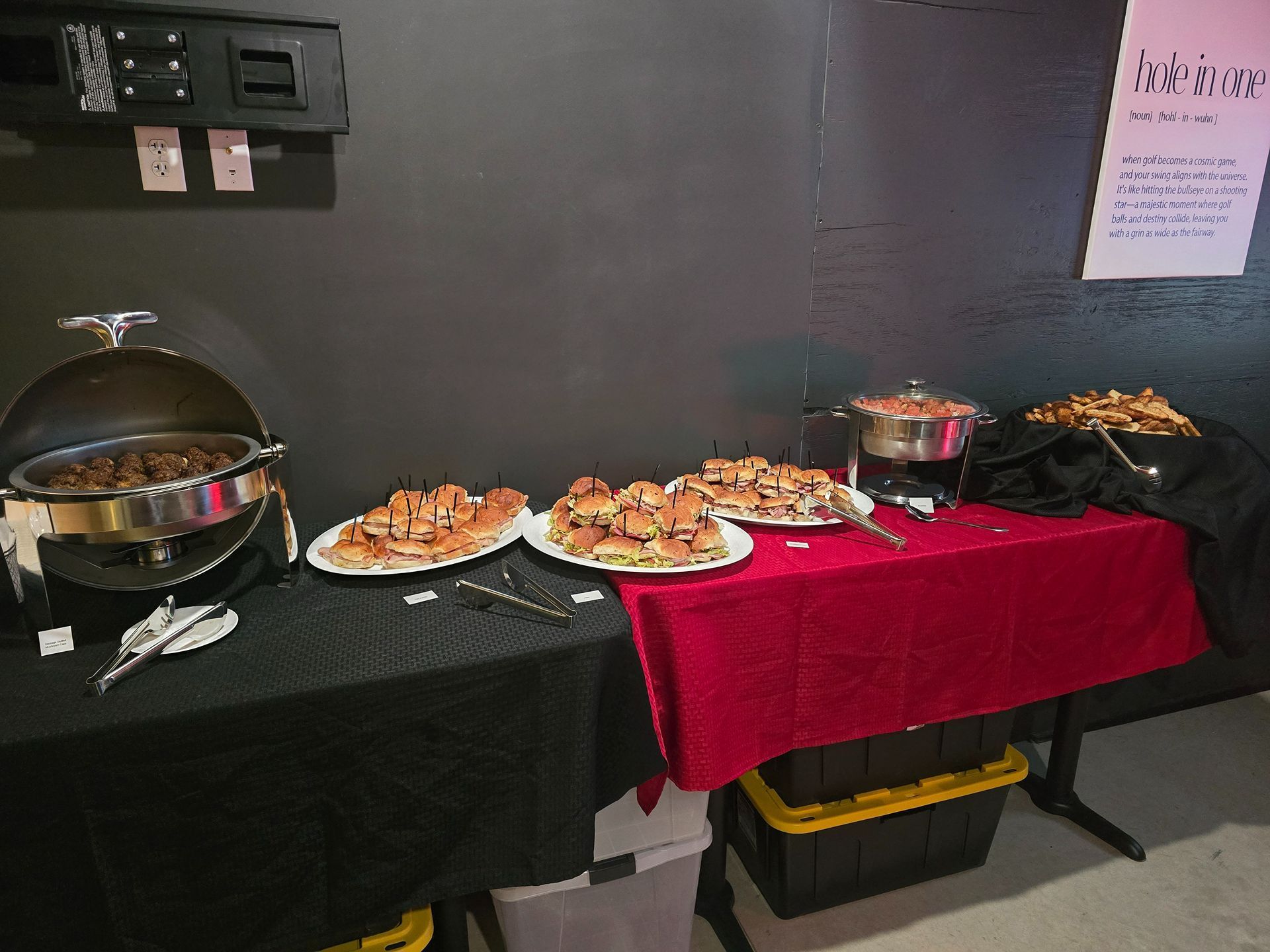 Buffet table with serving dishes and plates of food on black and red tablecloths against a dark wall.