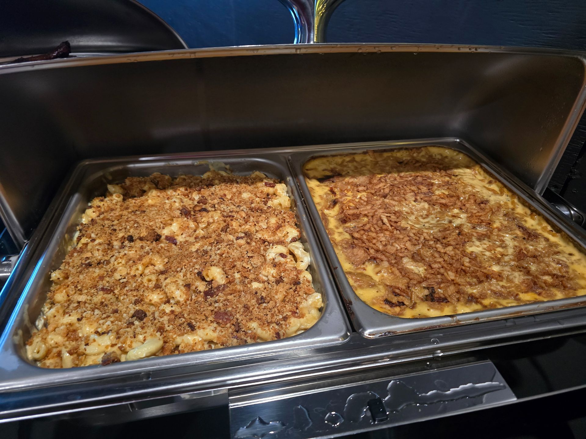Two trays of baked casserole under a buffet sneeze guard, topped with browned crumbs