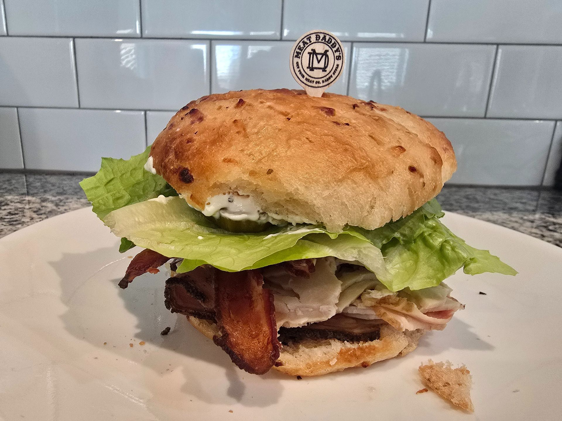 Tall sandwich with lettuce and bacon on a white plate against a tiled wall.