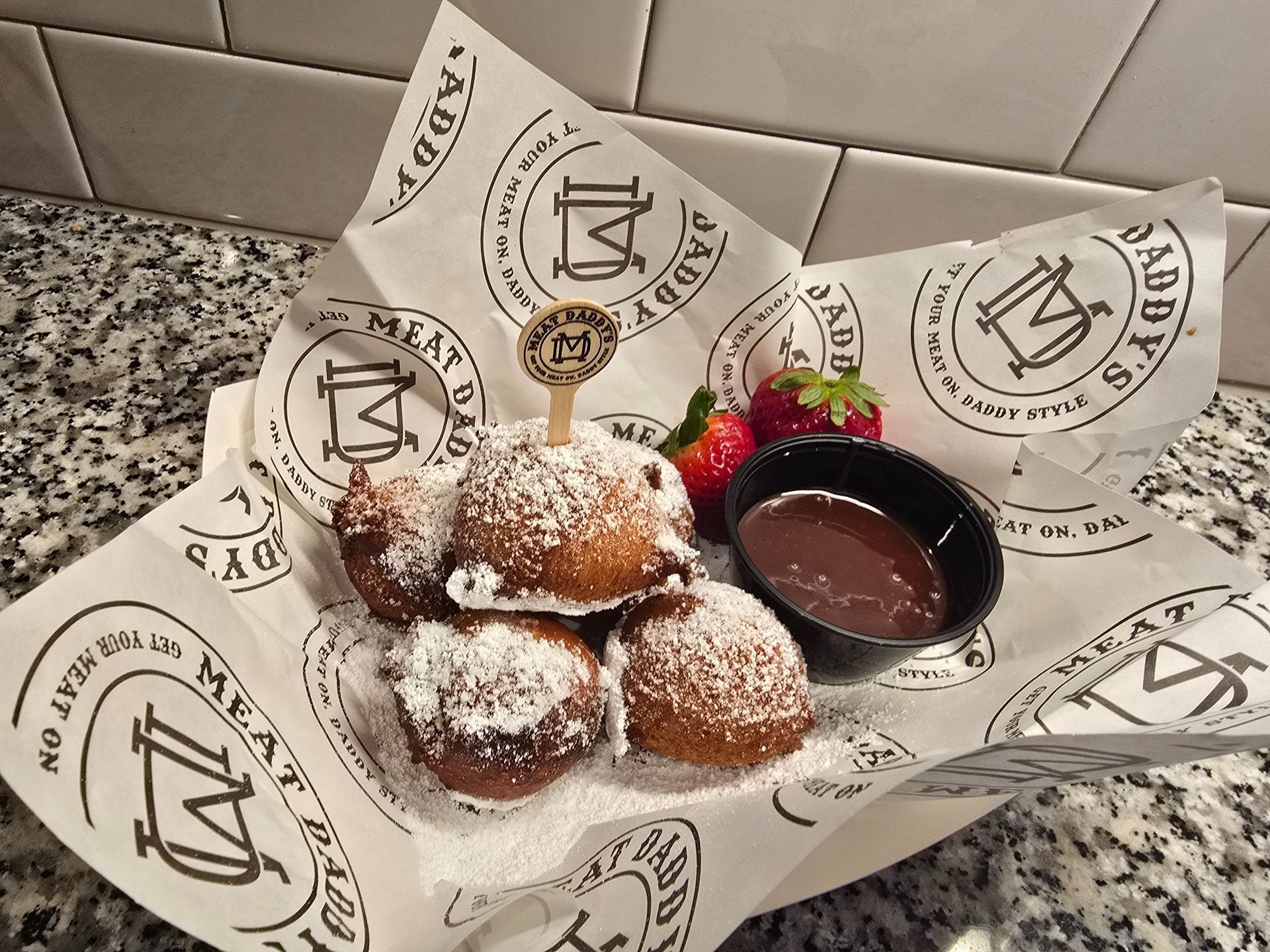 Dessert plate with powdered sugar doughnut bites, chocolate sauce, and a small garnish on branded paper