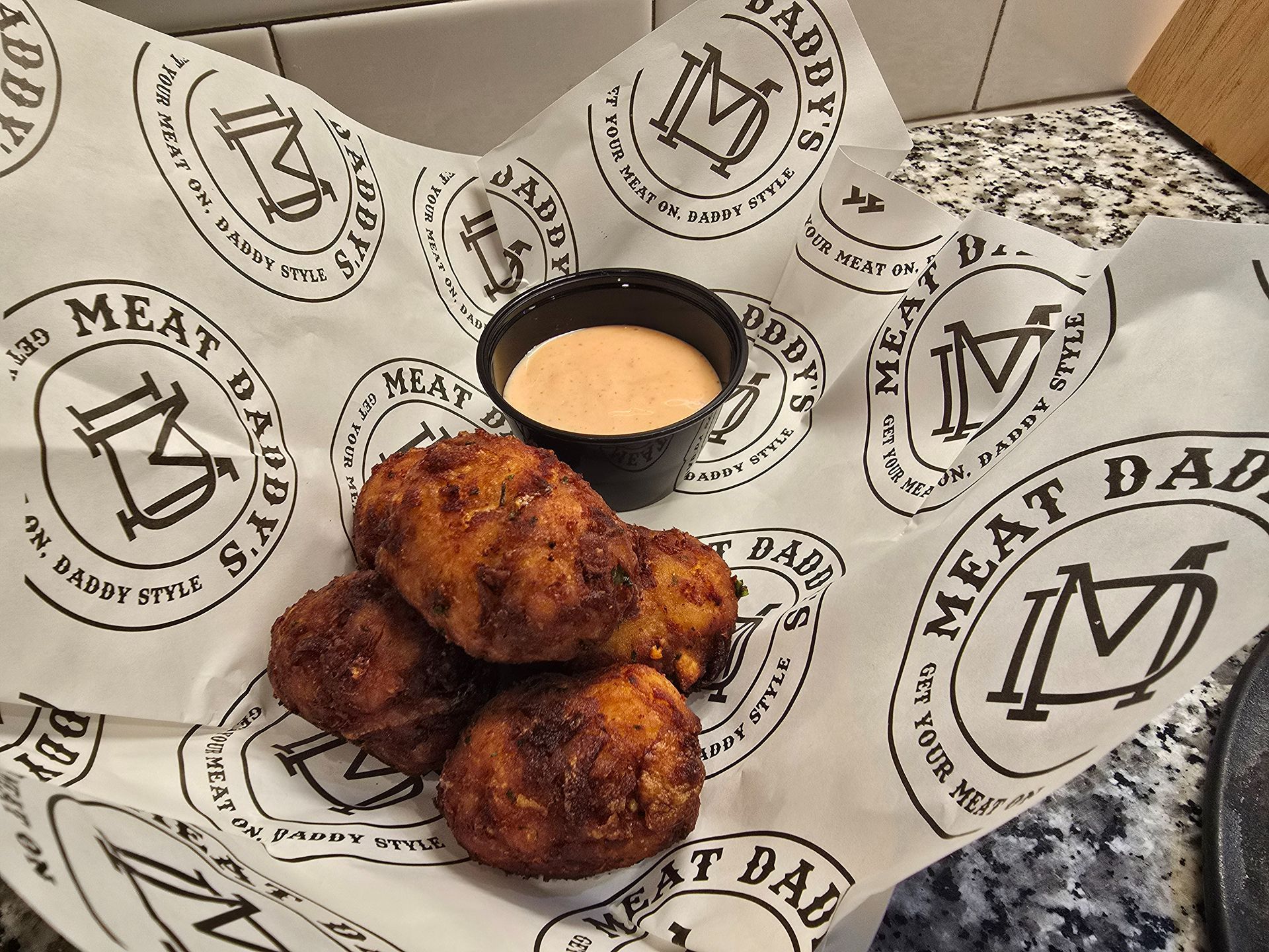 Spicy fried meatballs in branded paper, with a small cup of dipping sauce on a table