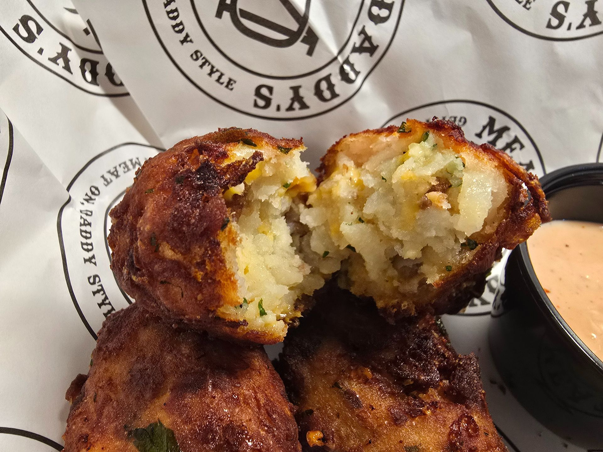 Golden-brown fried croquettes, one cut open to show a creamy mashed filling, on branded paper with dipping sauce