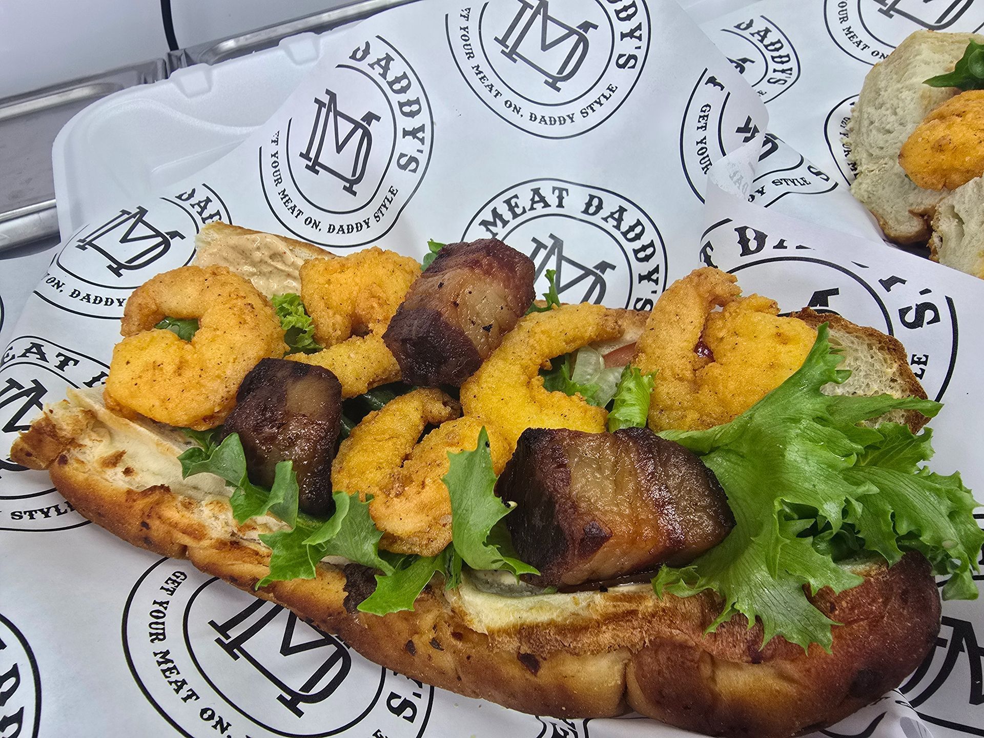 Grilled sandwich topped with lettuce, fried shrimp, and dark sauce on branded paper, with fries in the corner