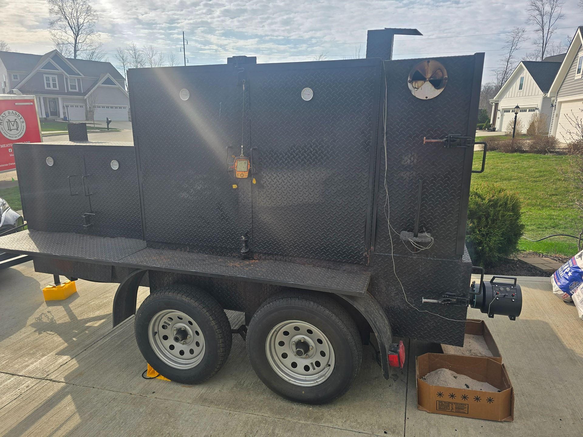 Black utility trailer with two wheels, hitched on a driveway with cardboard boxes and yellow wheel chocks.
