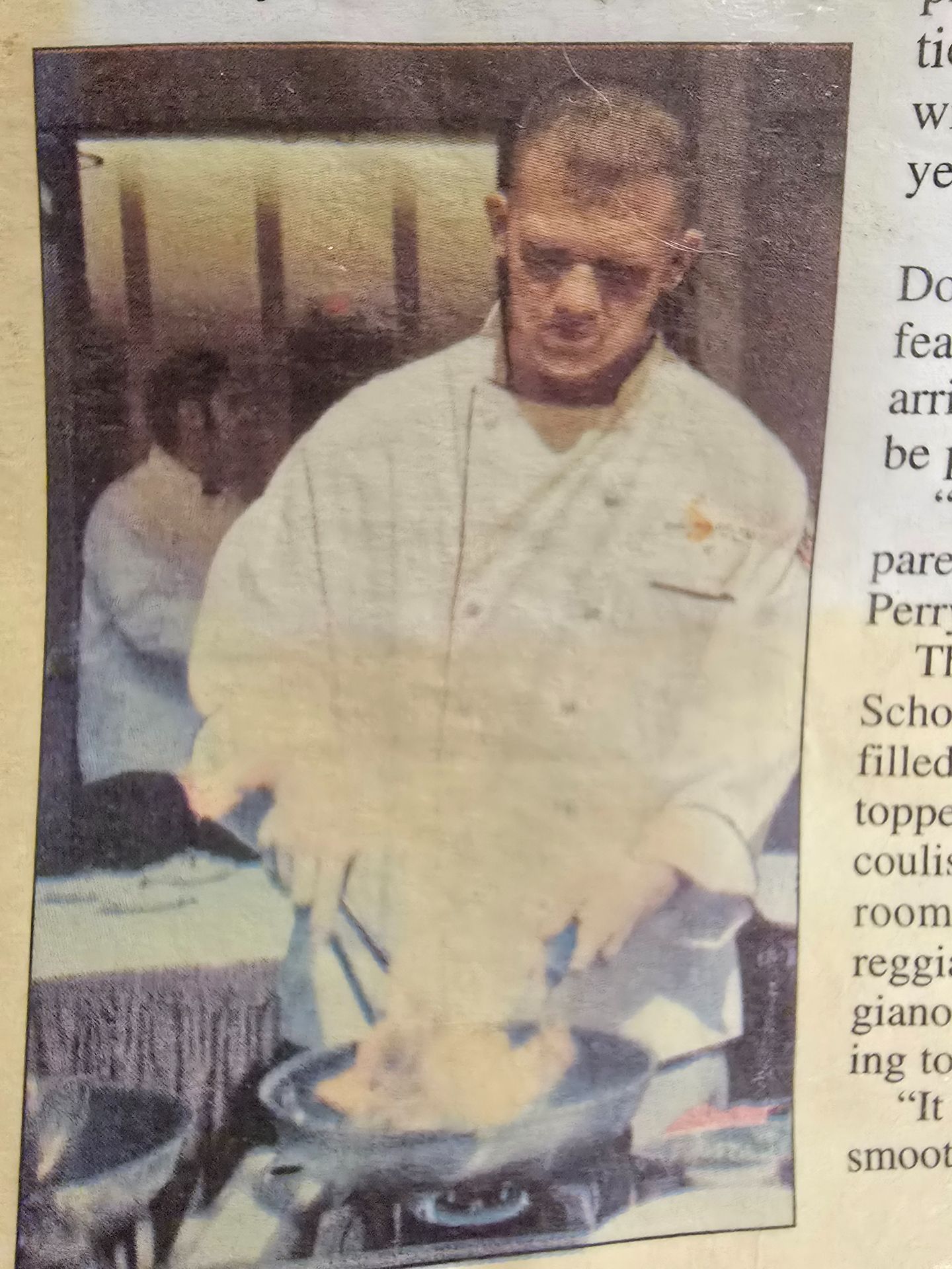 Man in a white jacket stirring food in a pan, standing at a stove in a kitchen.