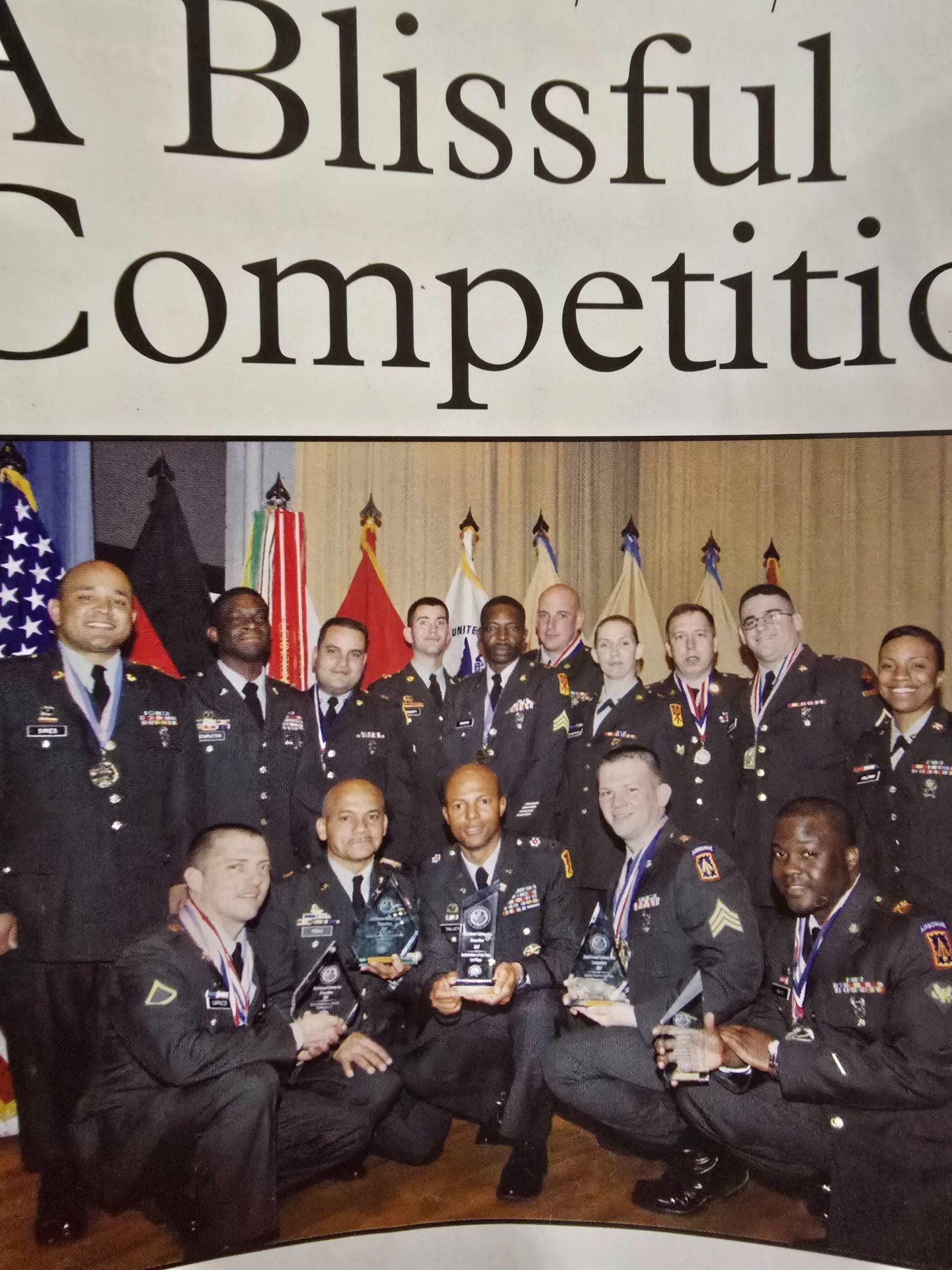Group photo of uniformed officials on a stage with flags and a banner reading “A Blissful Competitio”