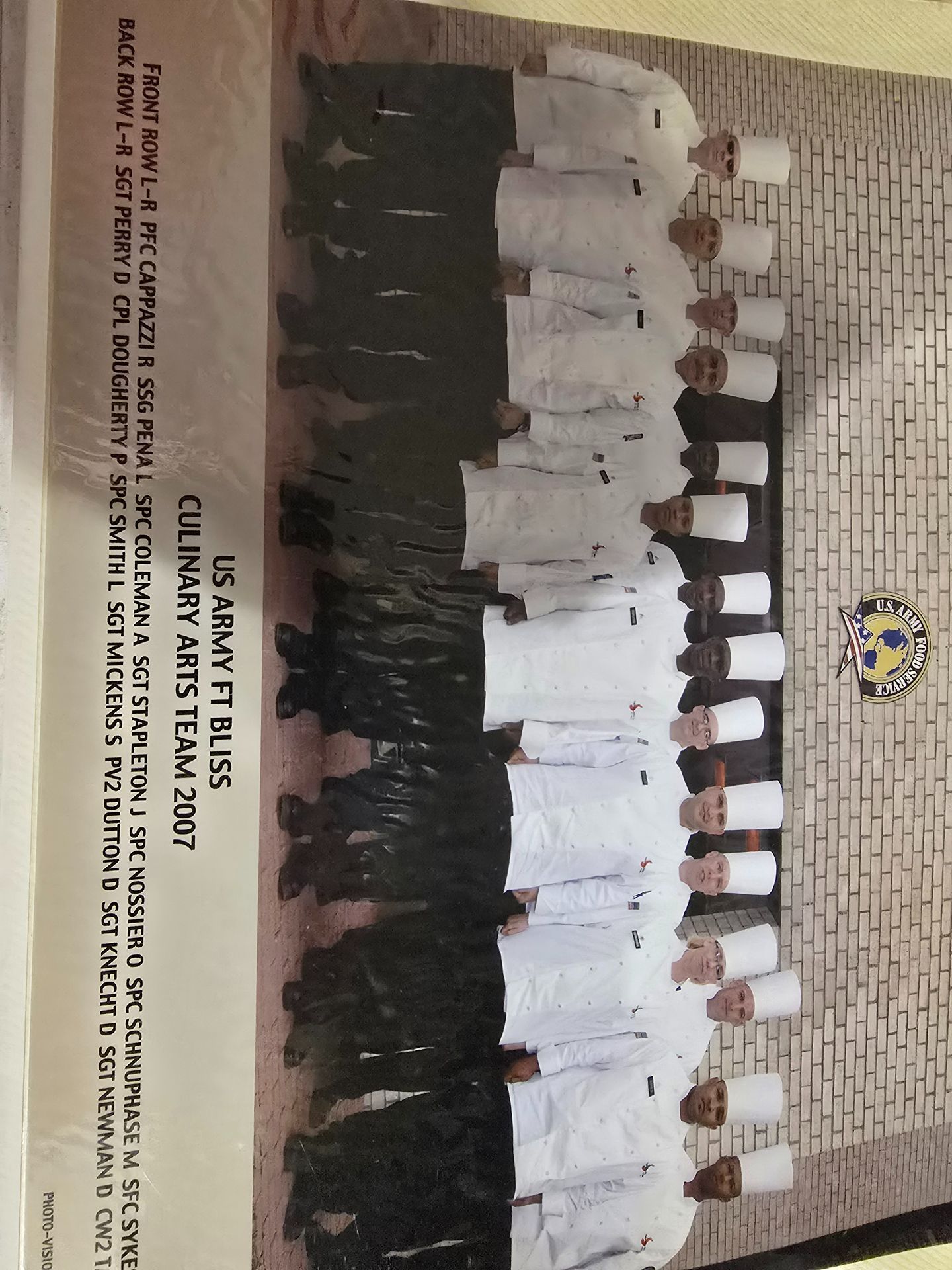 Group of people in white uniforms posing in front of a brick wall and school emblem during a formal event