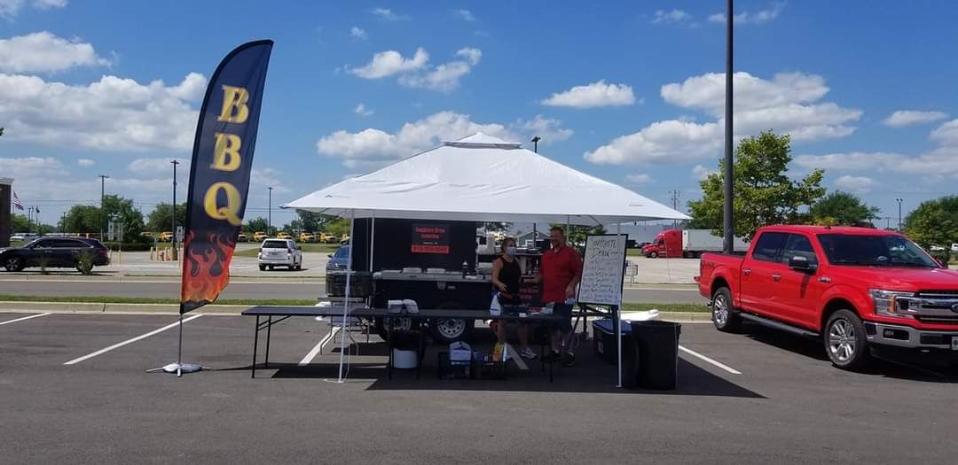 BBQ tent at a parking lot with a grill setup, blue flag, and red pickup truck under a sunny sky