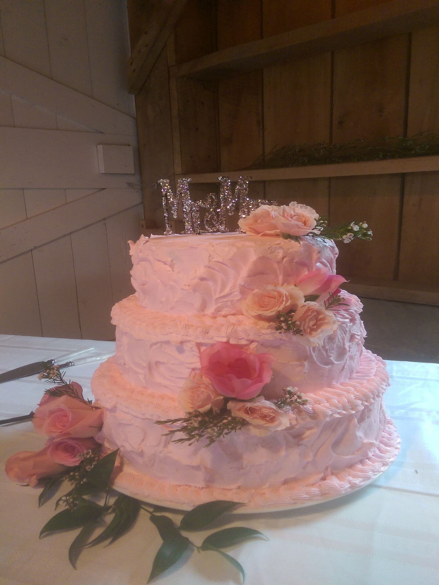 Pink three-tier wedding cake with ruffled icing and flowers on a table near a wooden wall