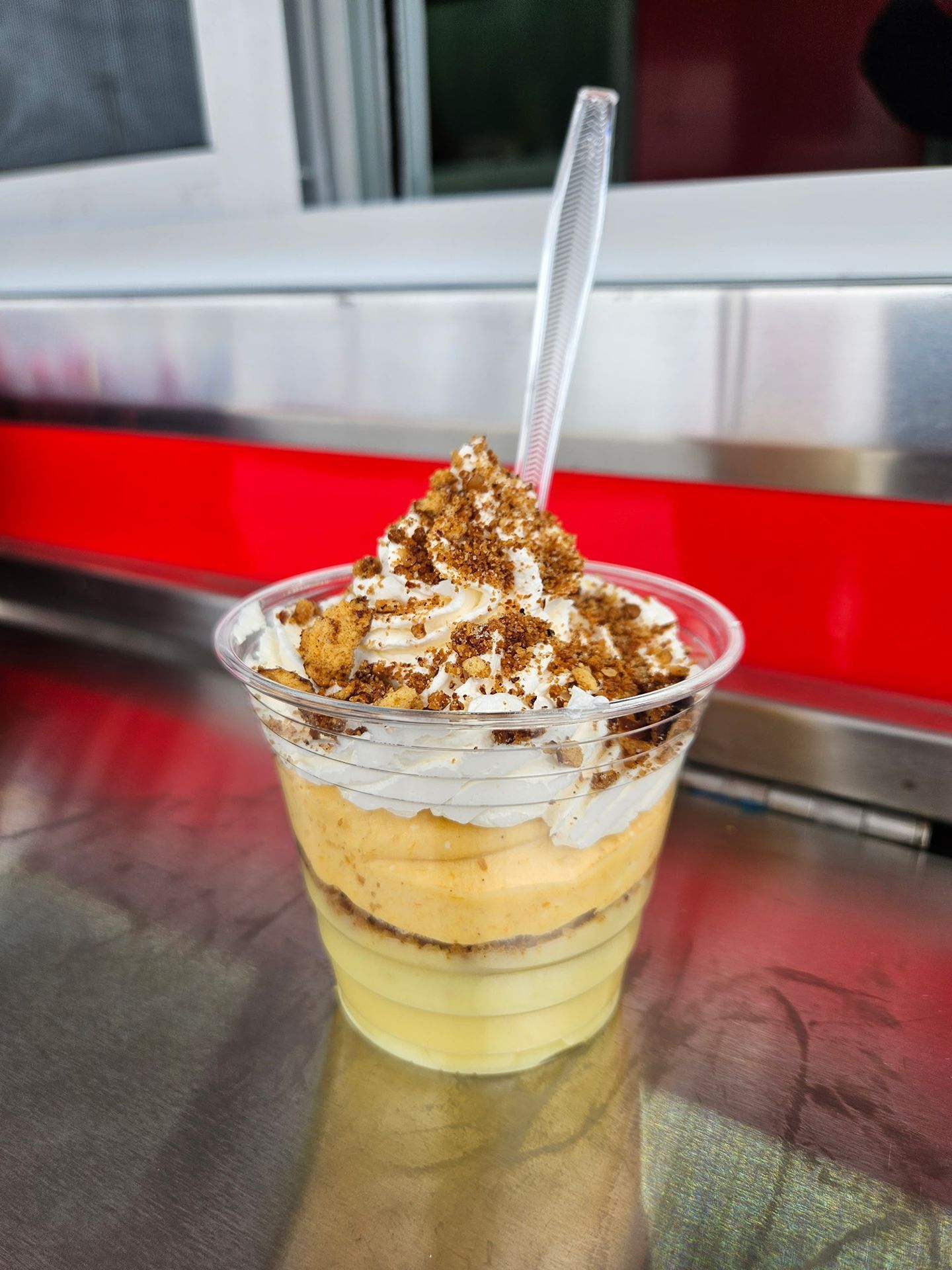 A layered dessert in a clear cup with whipped cream, caramel, and a spoon on a counter