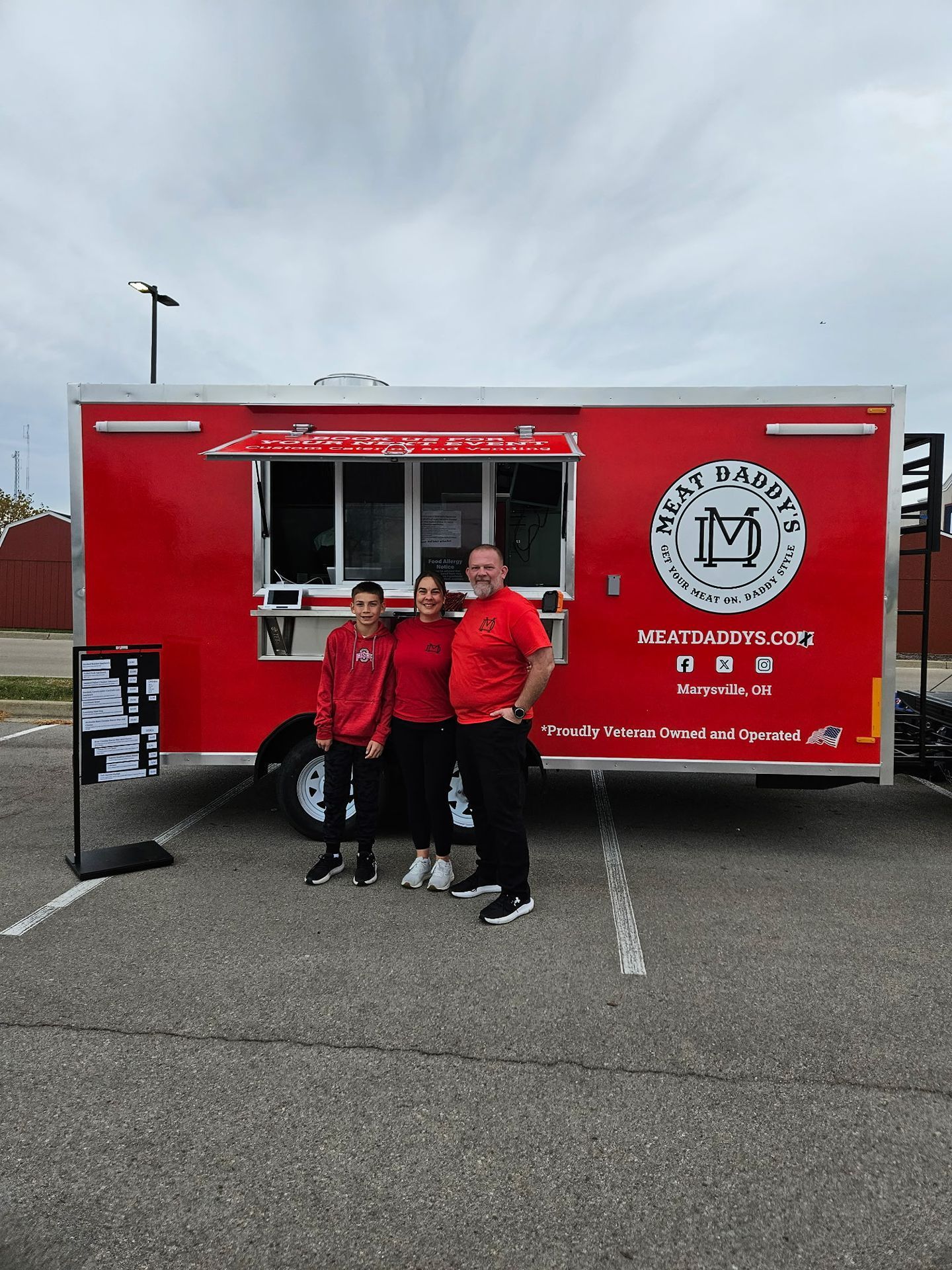 Three people in red jackets pose beside a red food truck in a parking lot.