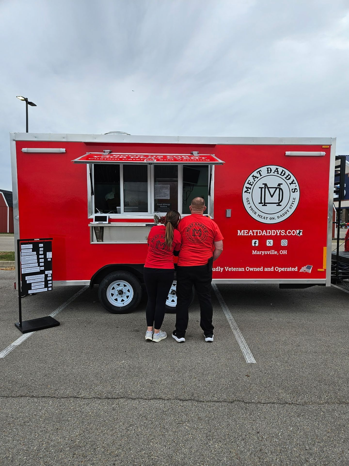 Red food truck with serving window and two people standing at the counter in a parking lot