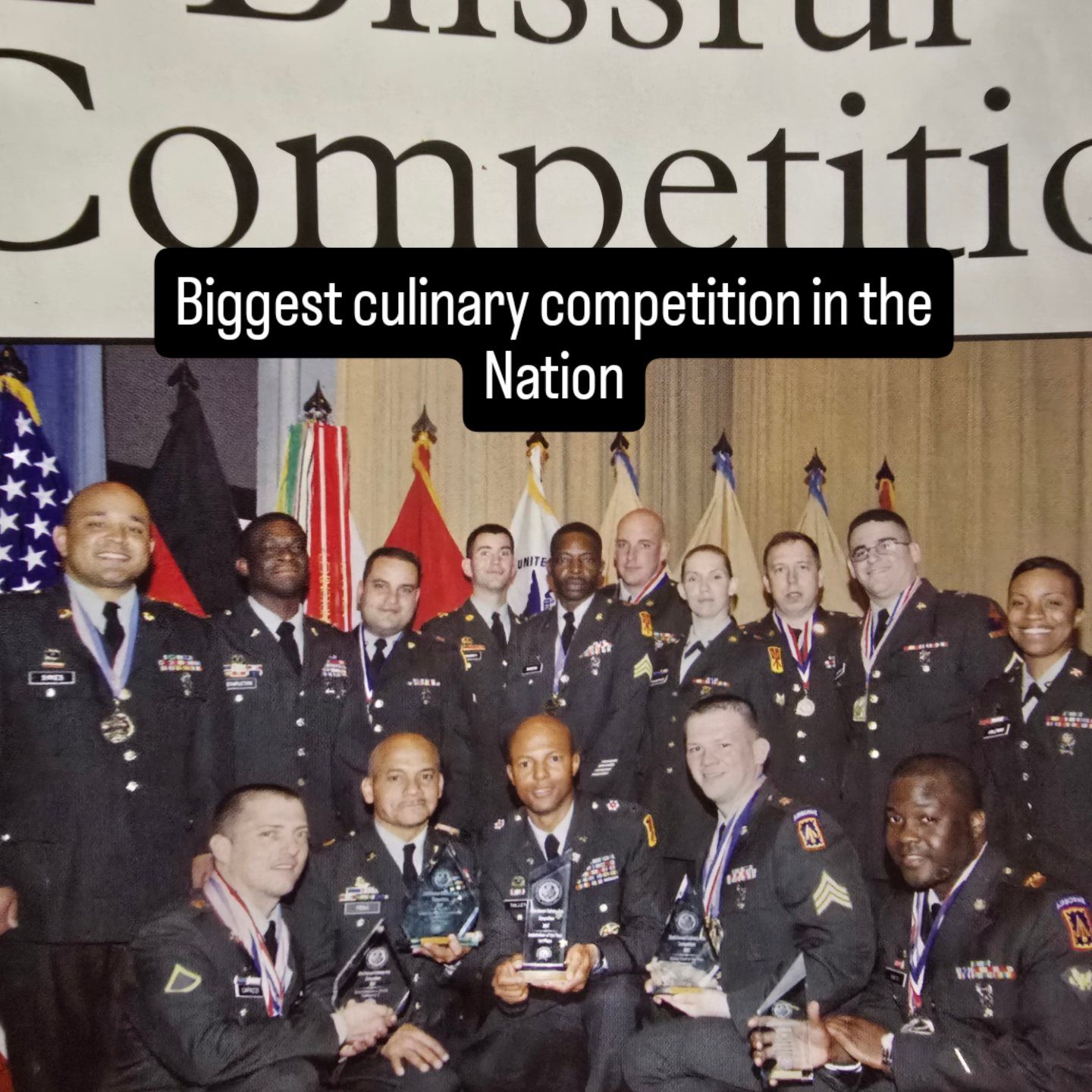 Group of uniformed culinary competition winners posing on stage with trophies and flags behind them