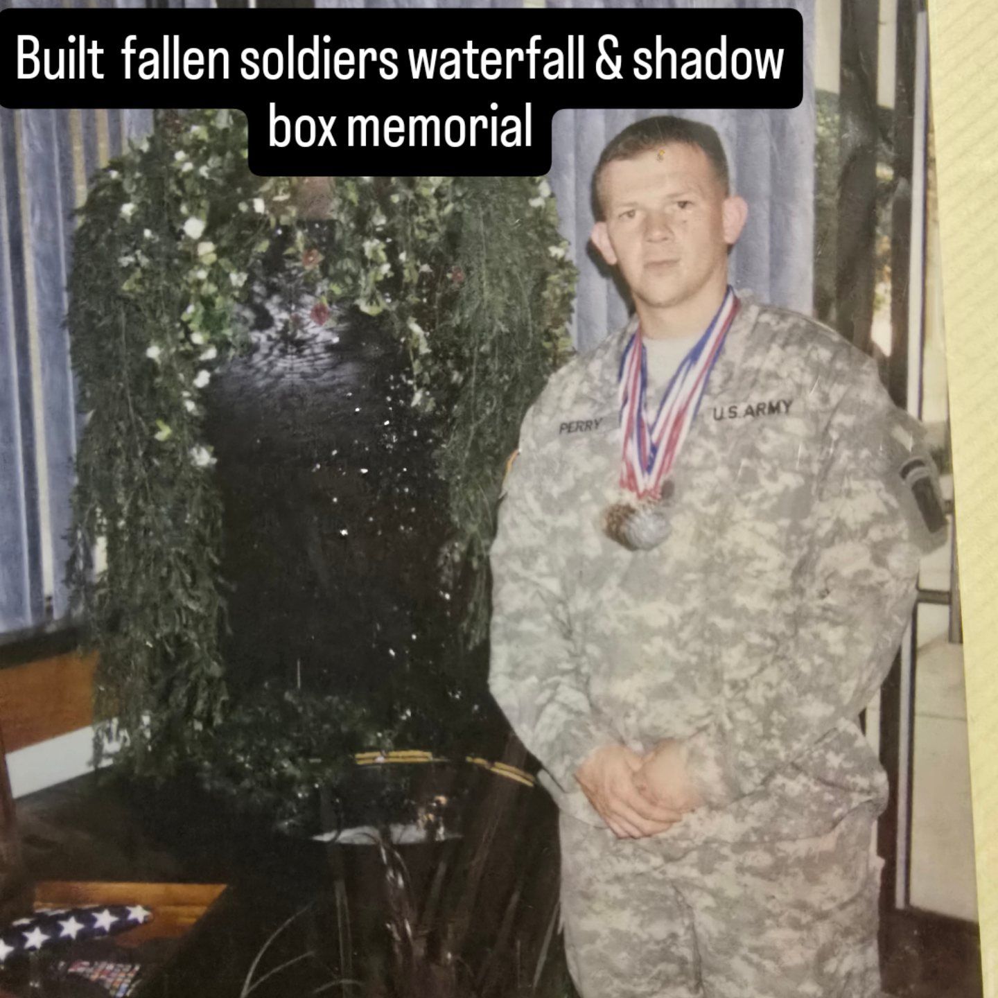 Soldier in camouflage stands beside a waterfall and shadow box memorial with medals displayed