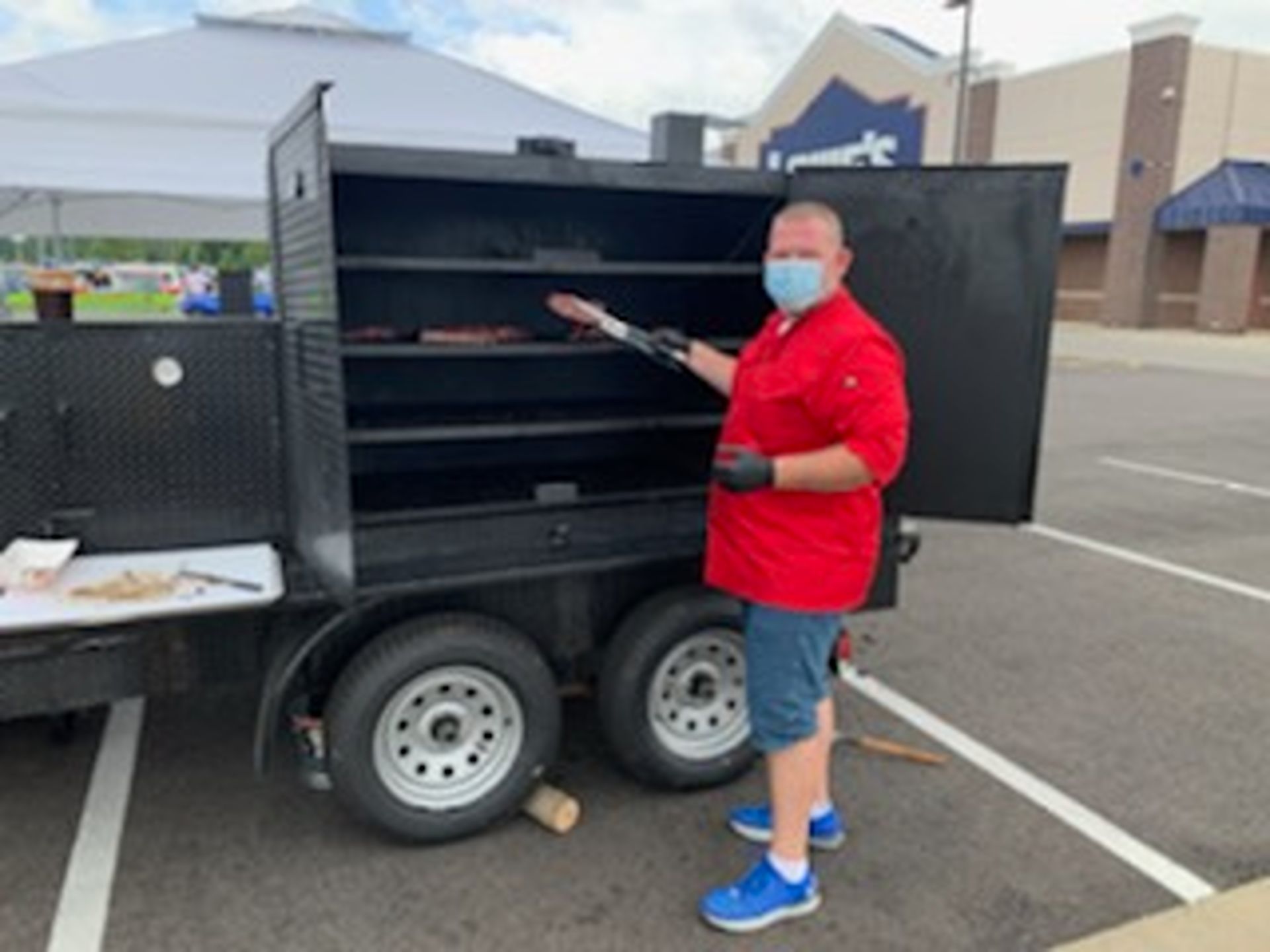 Man in red shirt and mask standing by a black dual-axle trailer grill in a parking lot