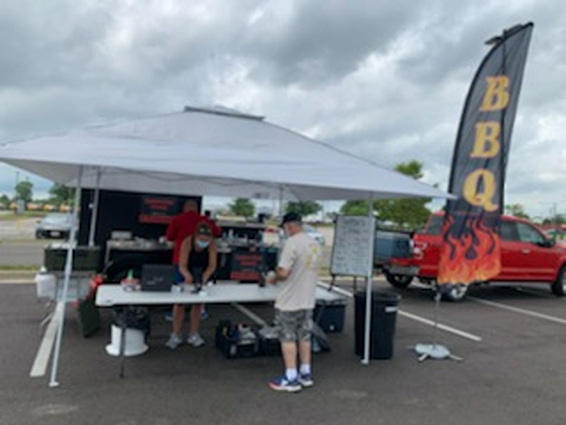 Outdoor BBQ food stand under a white canopy with a flame “BBQ” flag and a red truck nearby