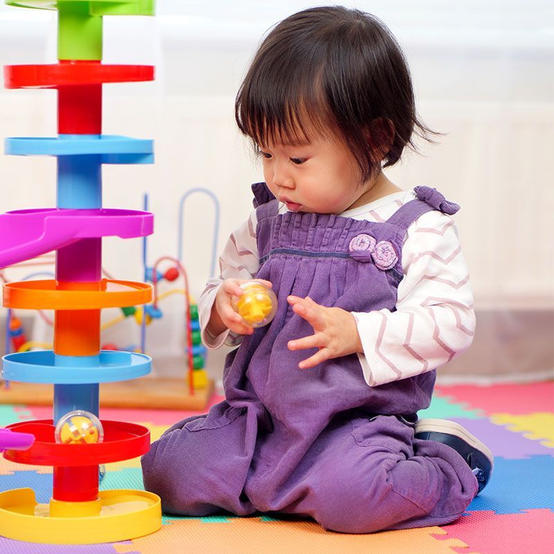 Toddler in purple overalls playing with a colorful toy tower on a play mat