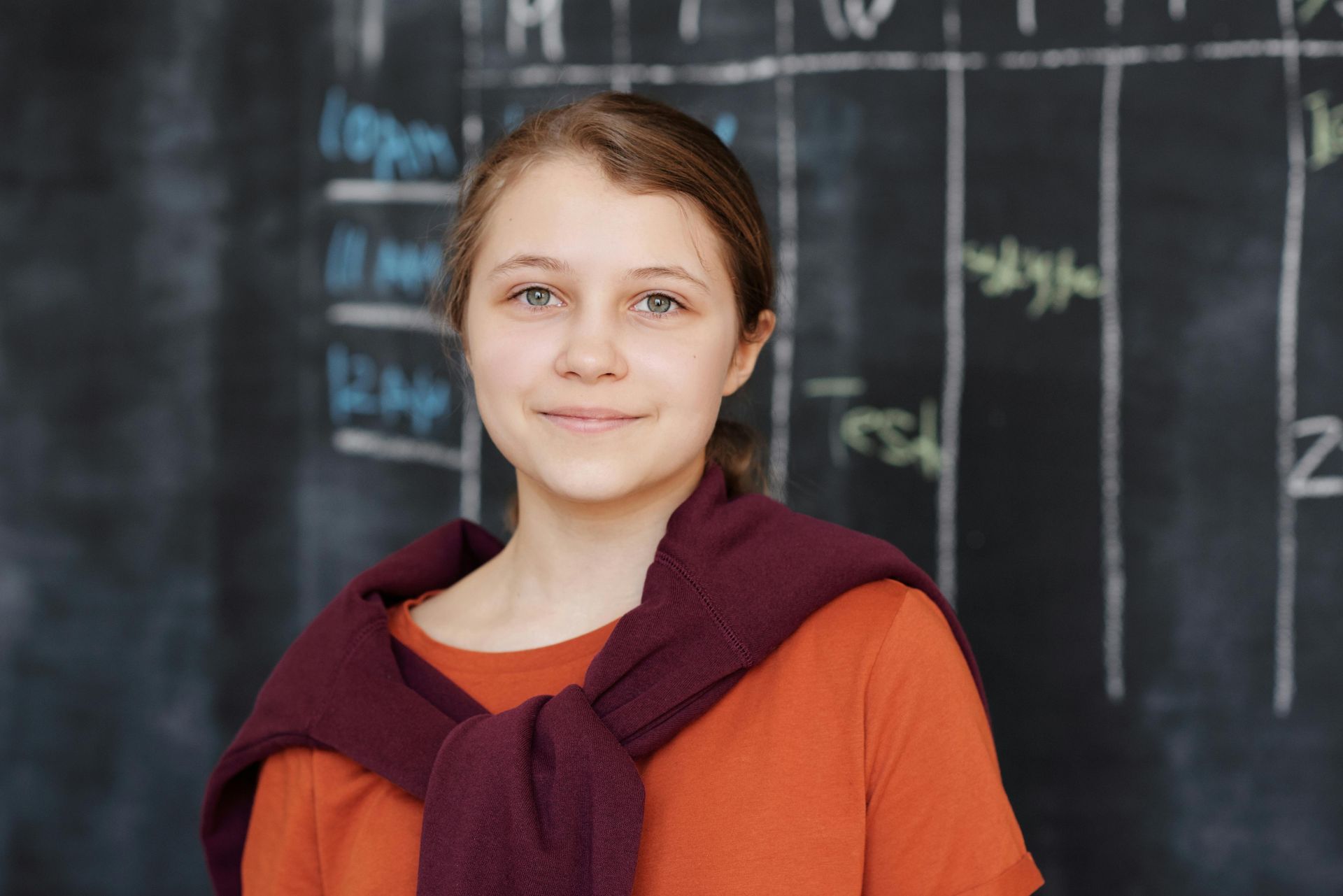 Young woman with a sweater around her shoulders smiles in front of a chalkboard.
