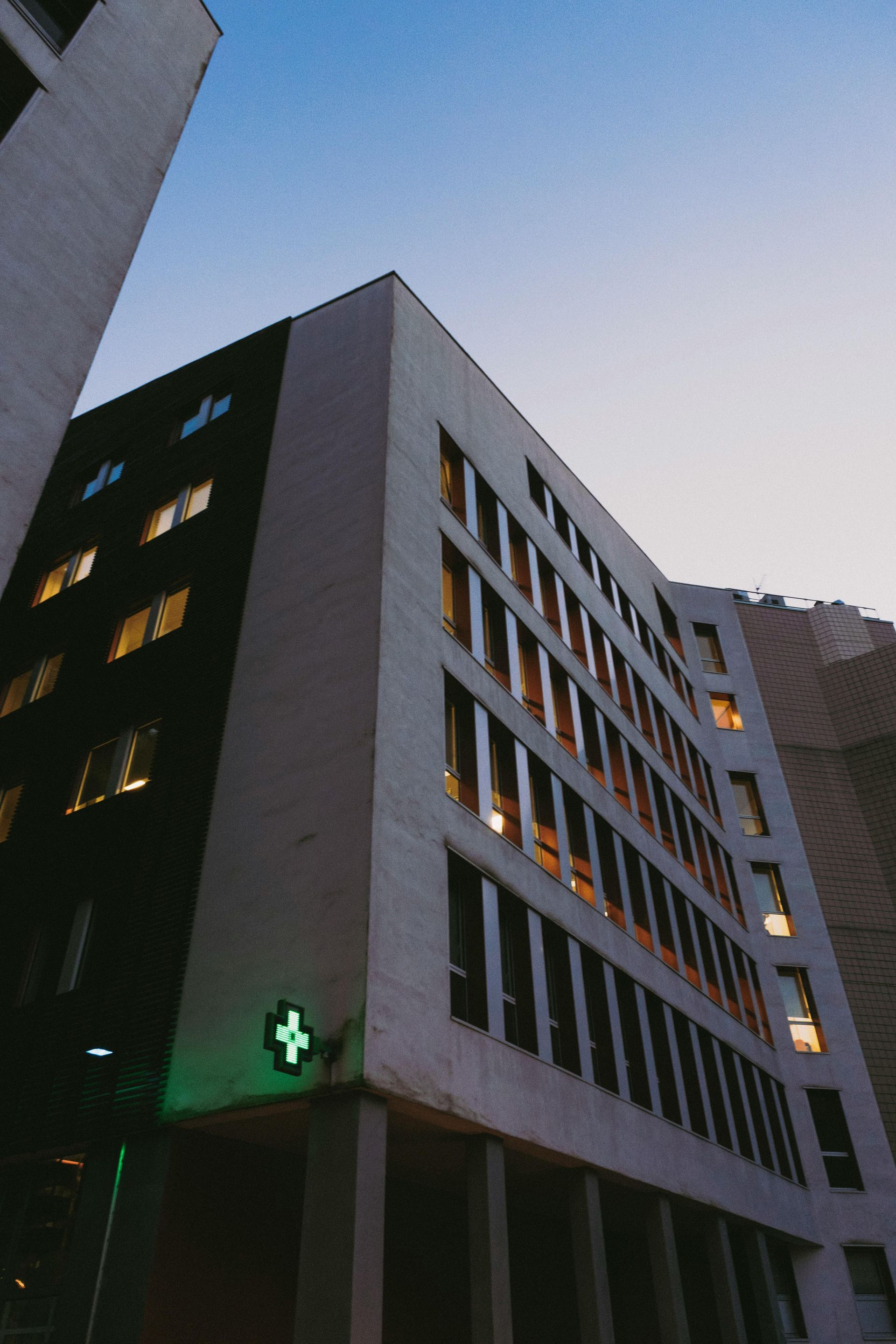 Modern hospital building at dusk with lit windows and a green cross sign
