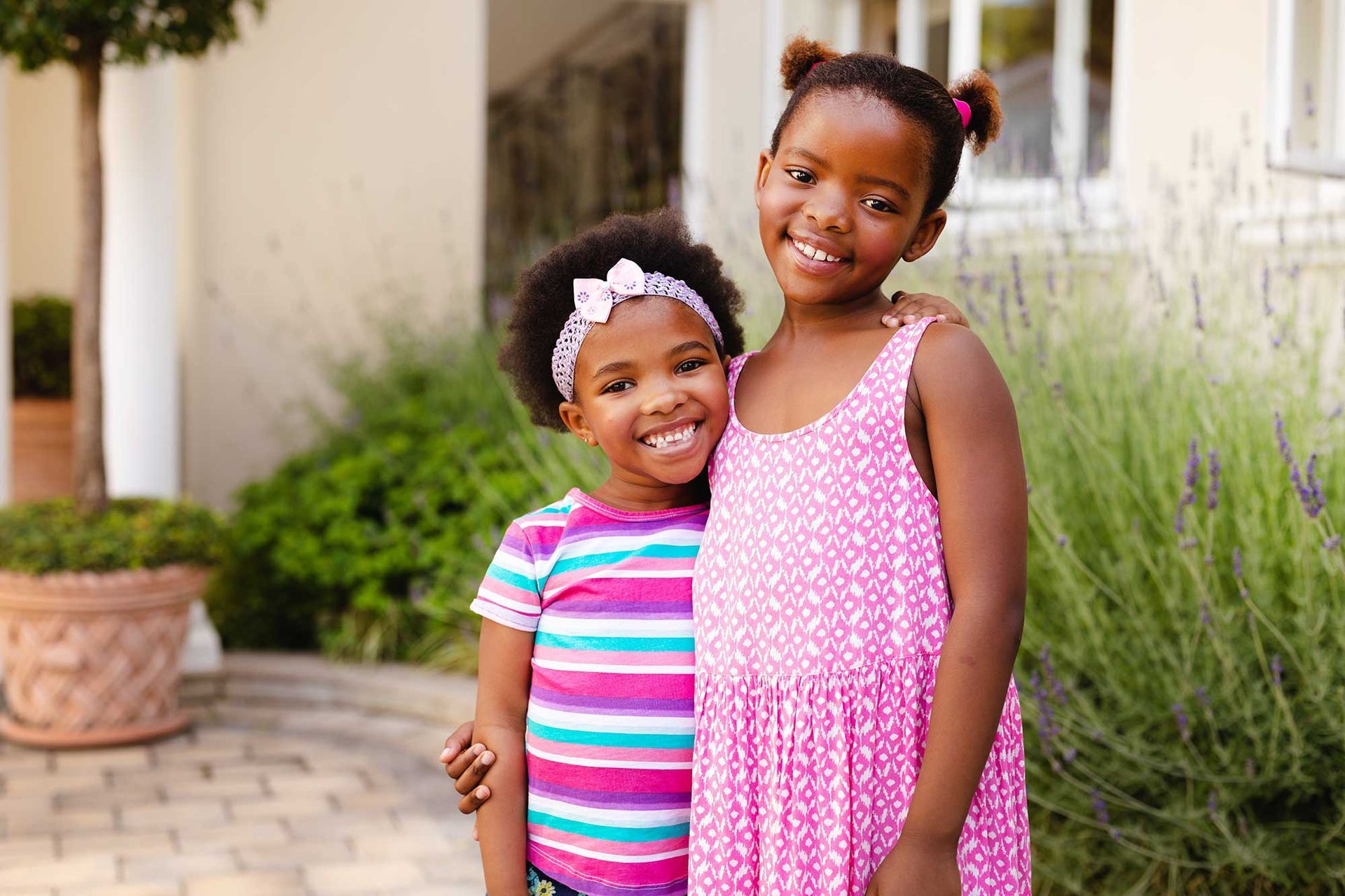 Two children smiling outdoors, standing on a brick walkway