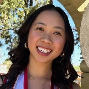 Woman with dark hair smiles, wearing a graduation sash and earrings, outdoors.