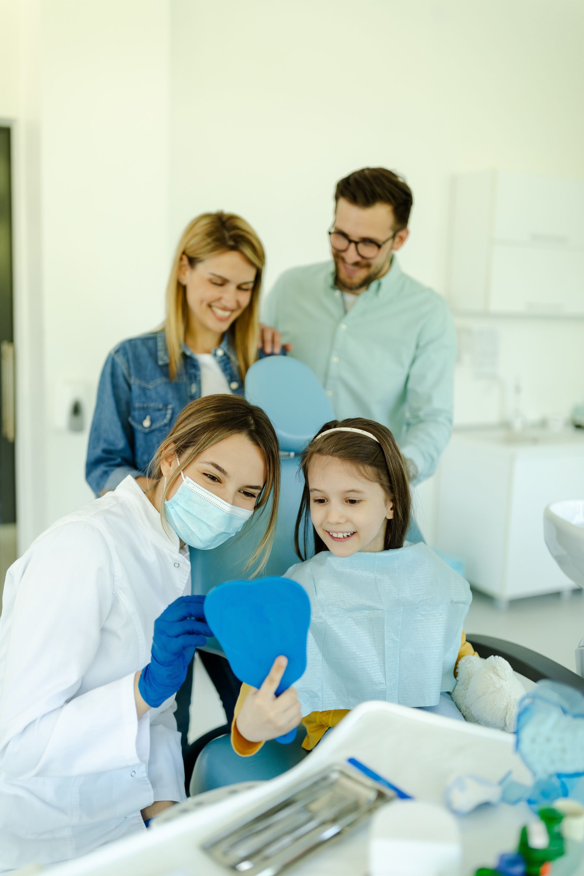 Dentist showing a young child her teeth in a mirror. Parents watch from behind. Bright dental office.