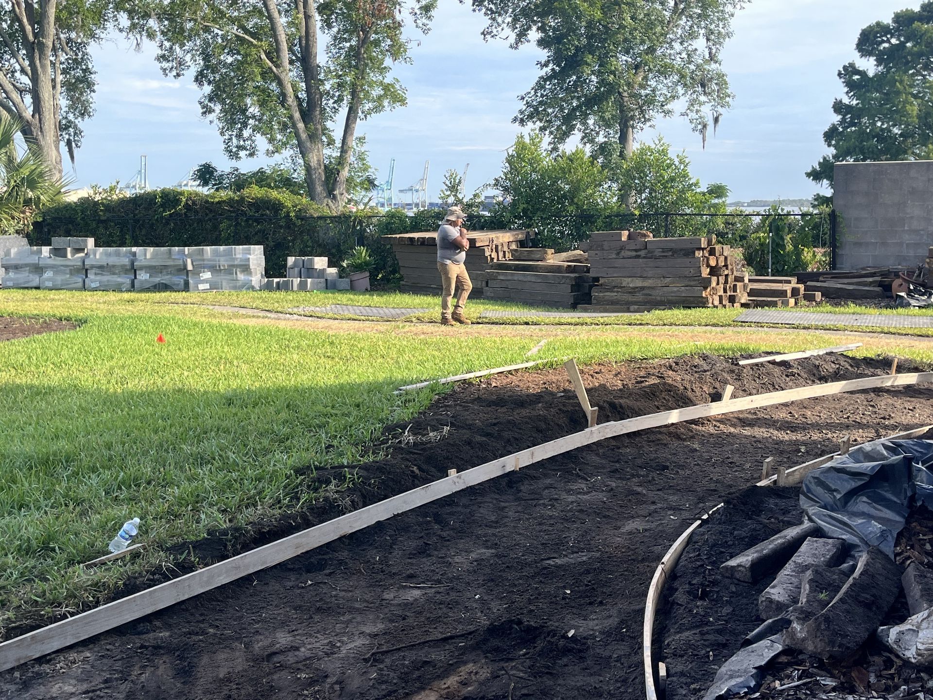 Person standing in a sunny backyard near stacked materials, with grass, trees, and a dirt path in the foreground.