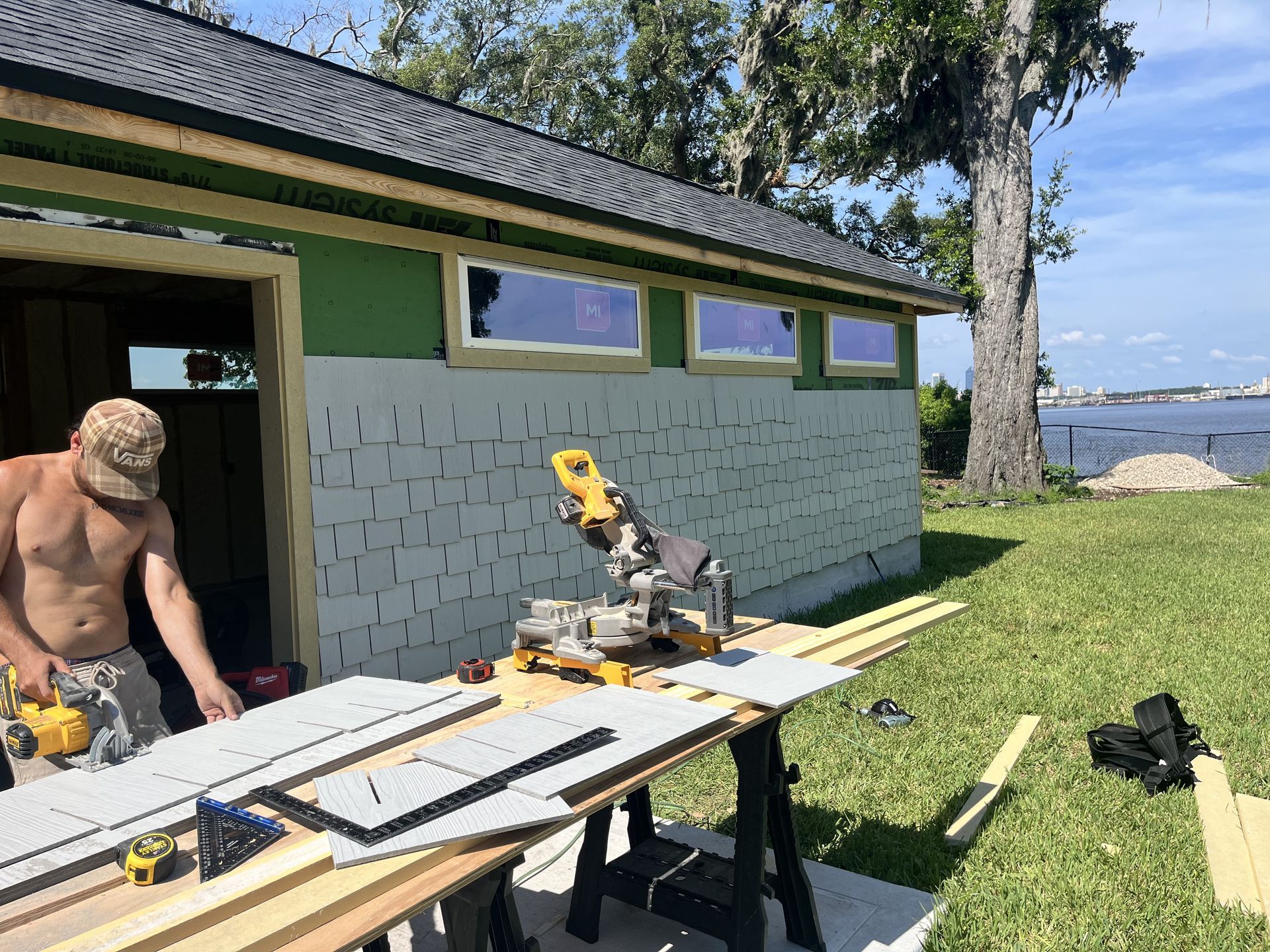 Person cutting boards outdoors beside a green lakeside shed with tools and lumber spread out on a table