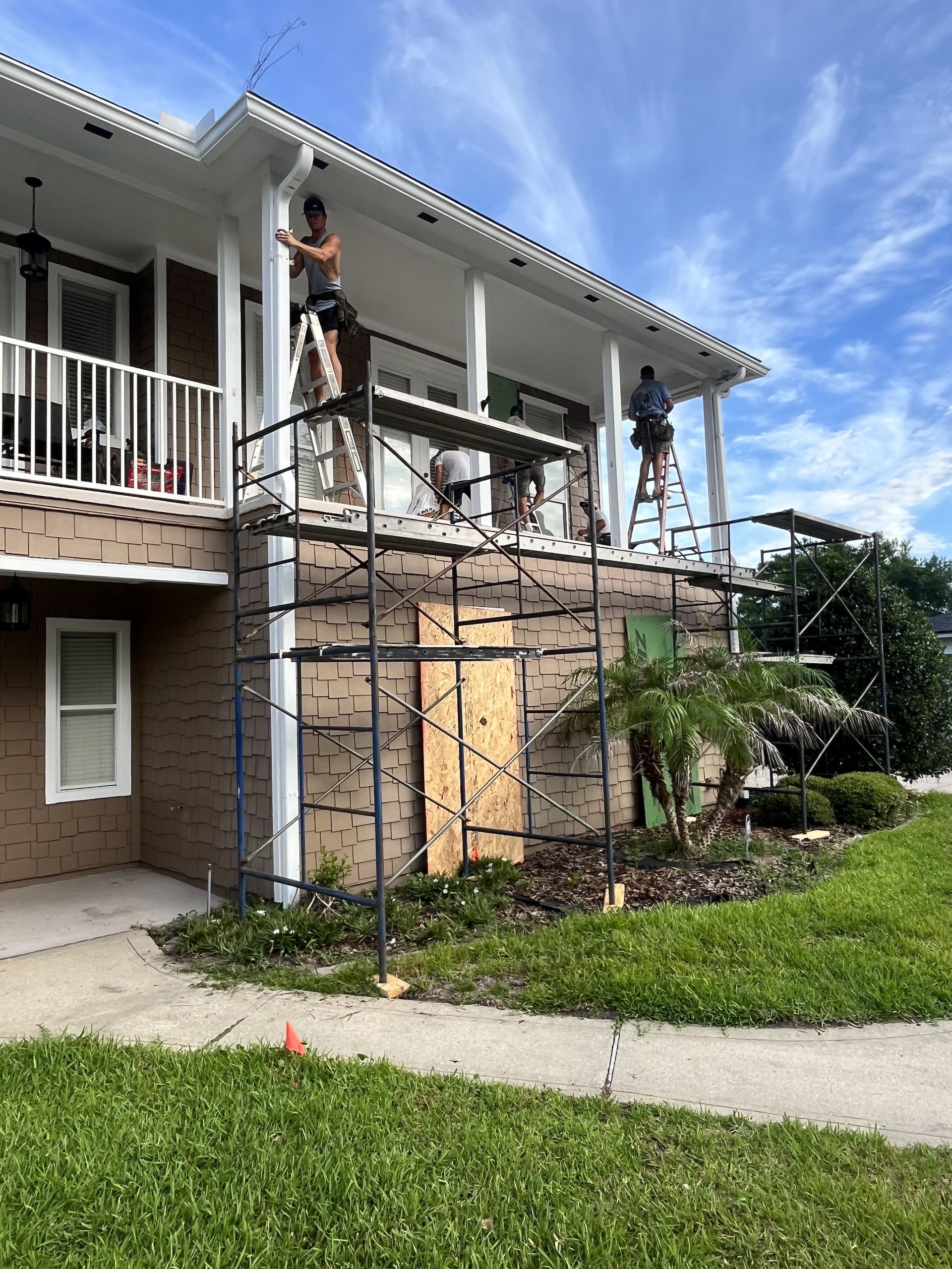 Two workers on scaffolding repairing the upper floor of a white house with a boarded-up balcony.