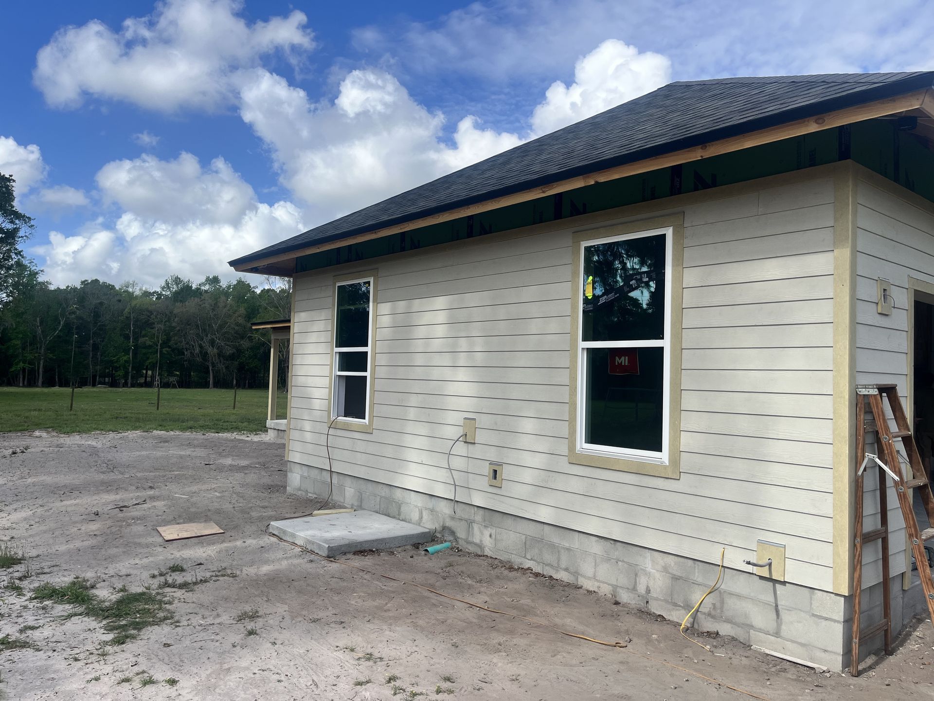 Small beige house under construction with dark roof, windows, and a dirt lot in front under a cloudy sky