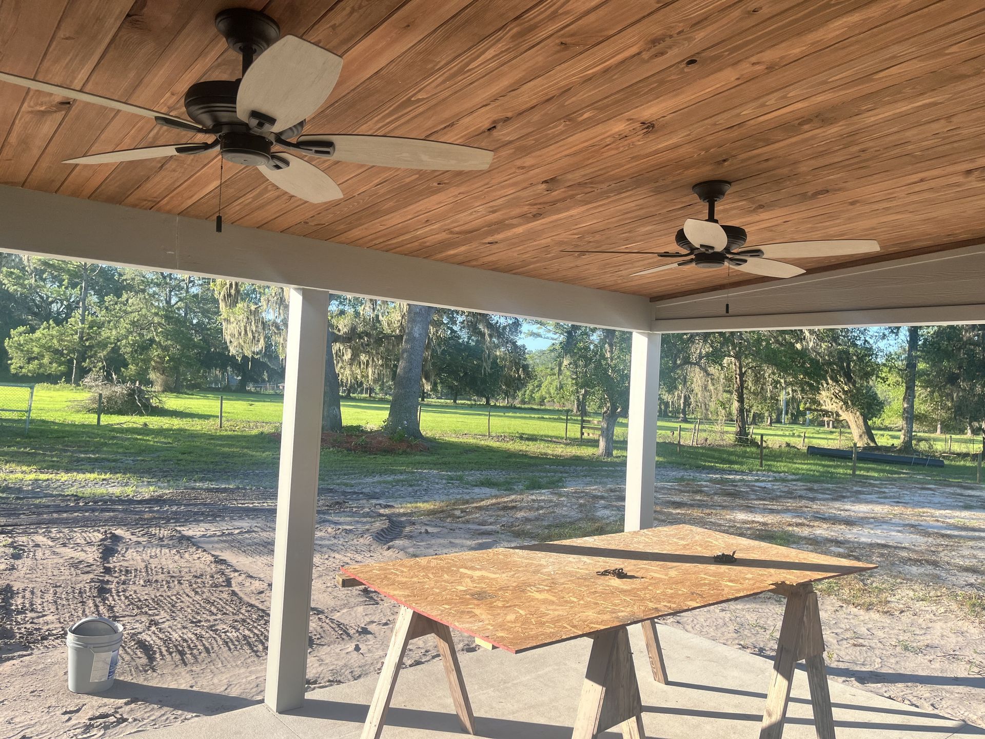 Covered outdoor patio with wood ceiling, two ceiling fans, and a folding table overlooking a grassy yard