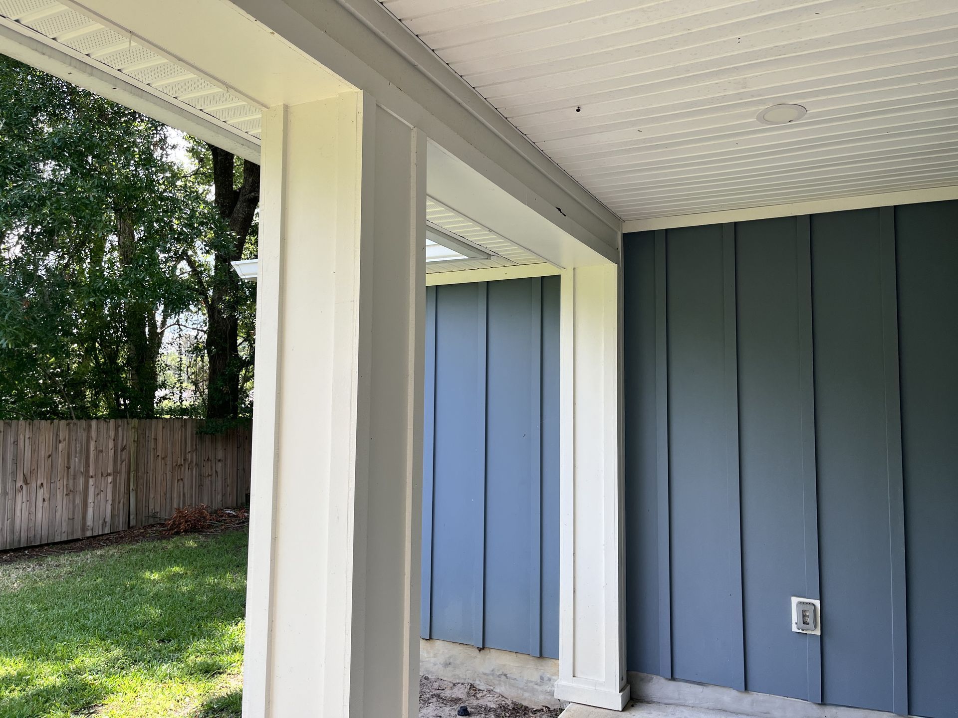 Covered patio with white posts, blue-gray wall, and grassy yard beyond a wooden fence.