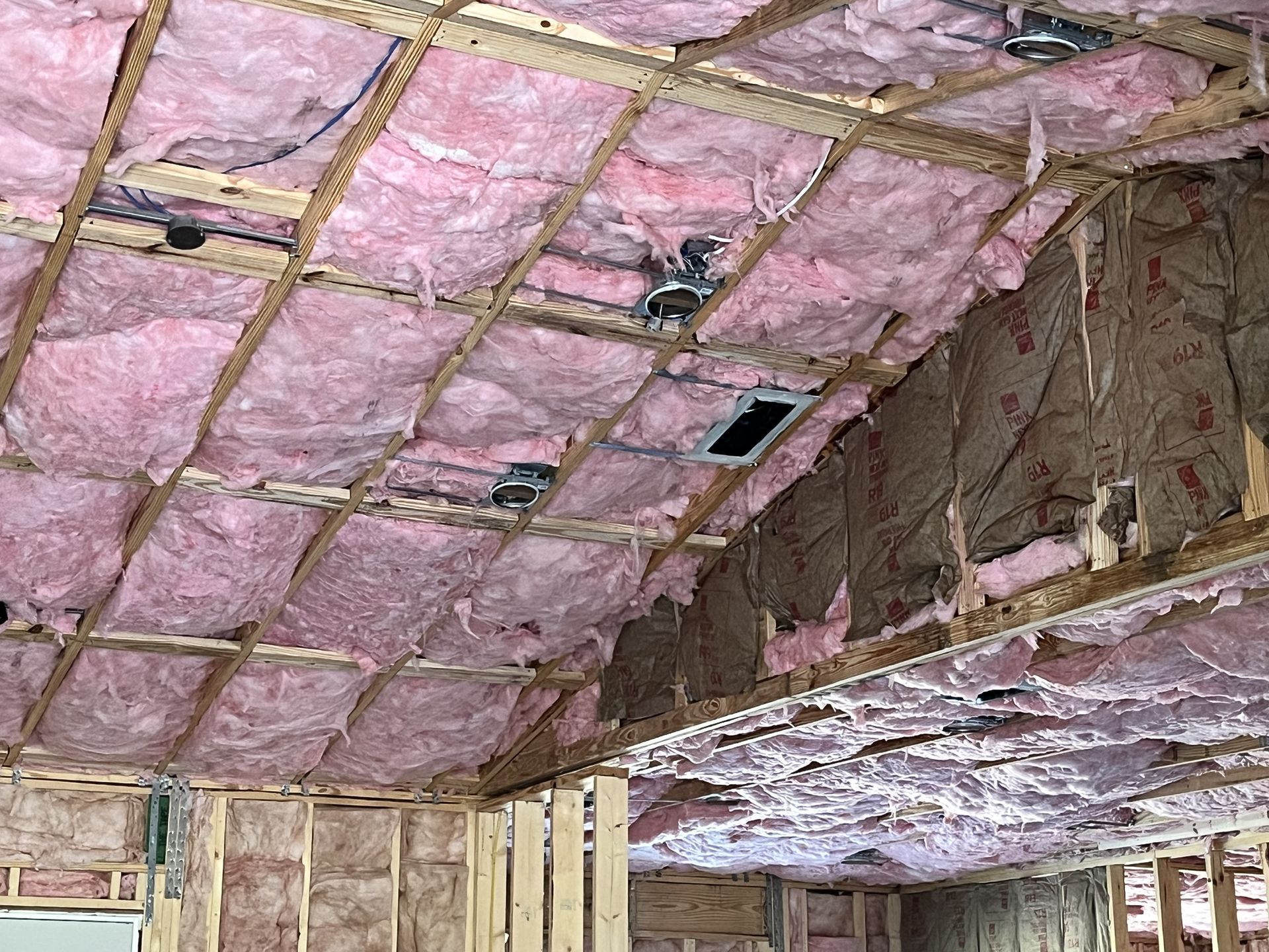 Pink insulation and exposed framing in an unfinished ceiling with a recessed light and vent opening.