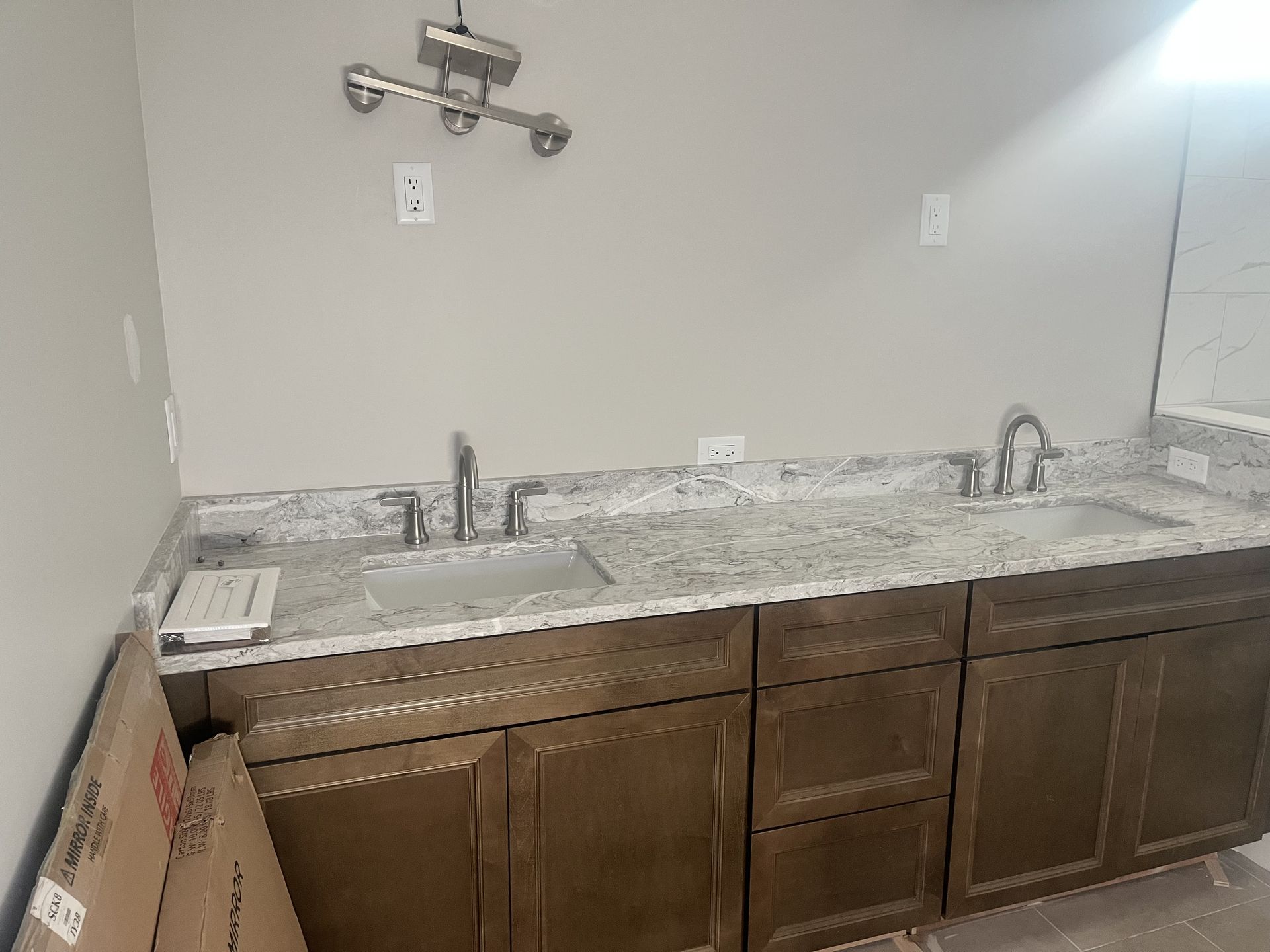 Unfinished bathroom vanity with marble countertop, double sink, and wall-mounted faucets above dark wood cabinets.