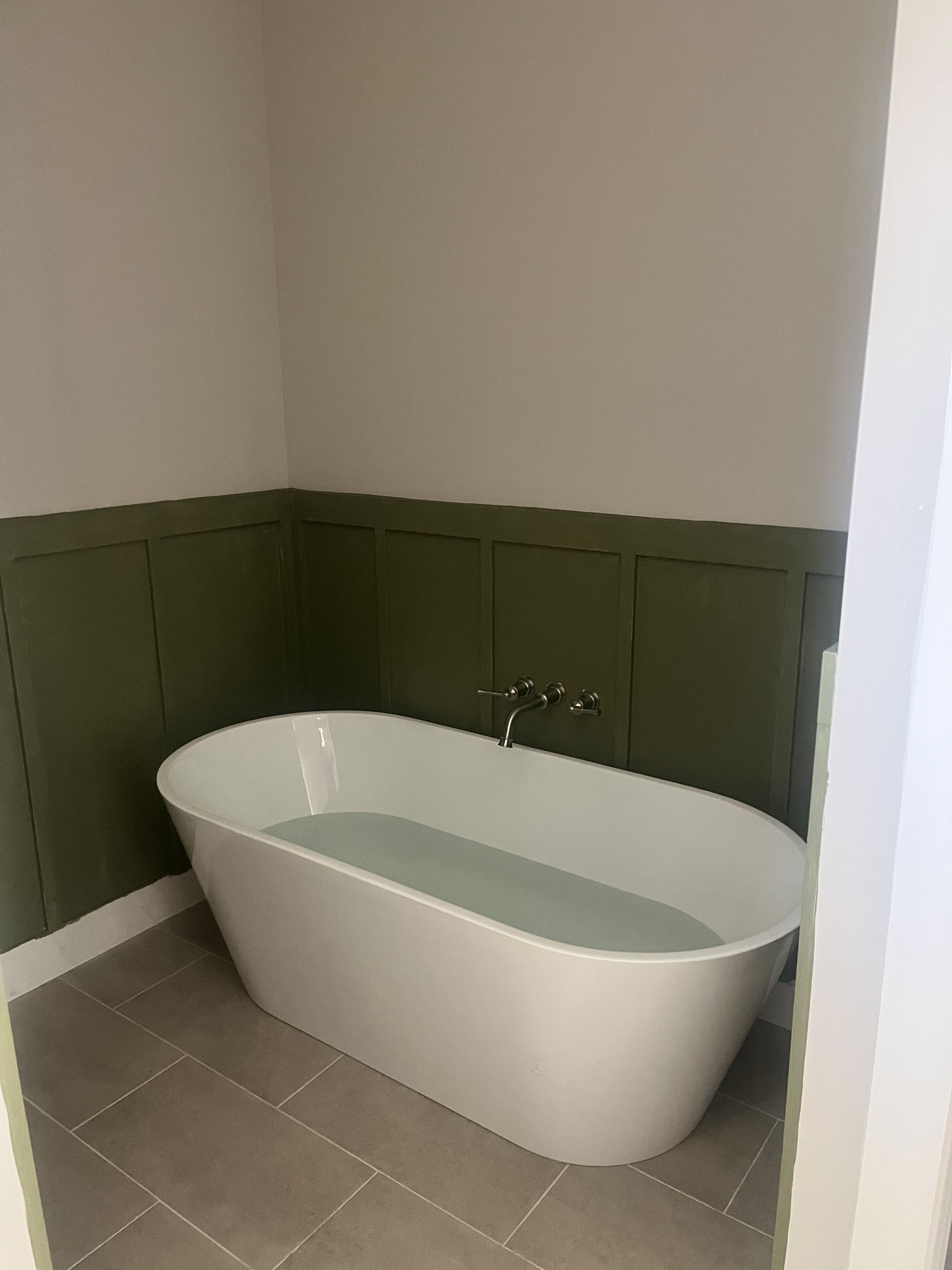 White freestanding bathtub in a small corner bathroom with green wainscoting and gray tile floor