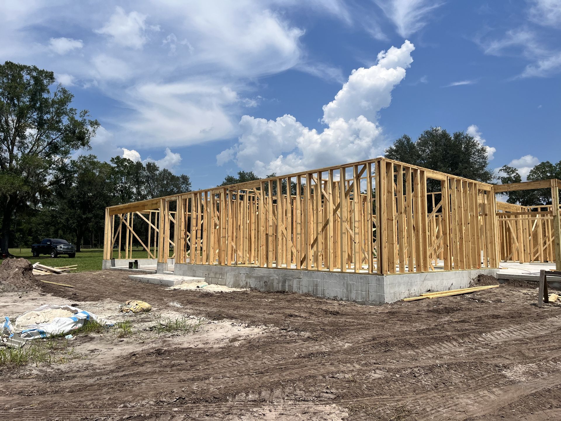 Wood-framed house under construction on a concrete foundation, with muddy ground and trees under a blue sky.
