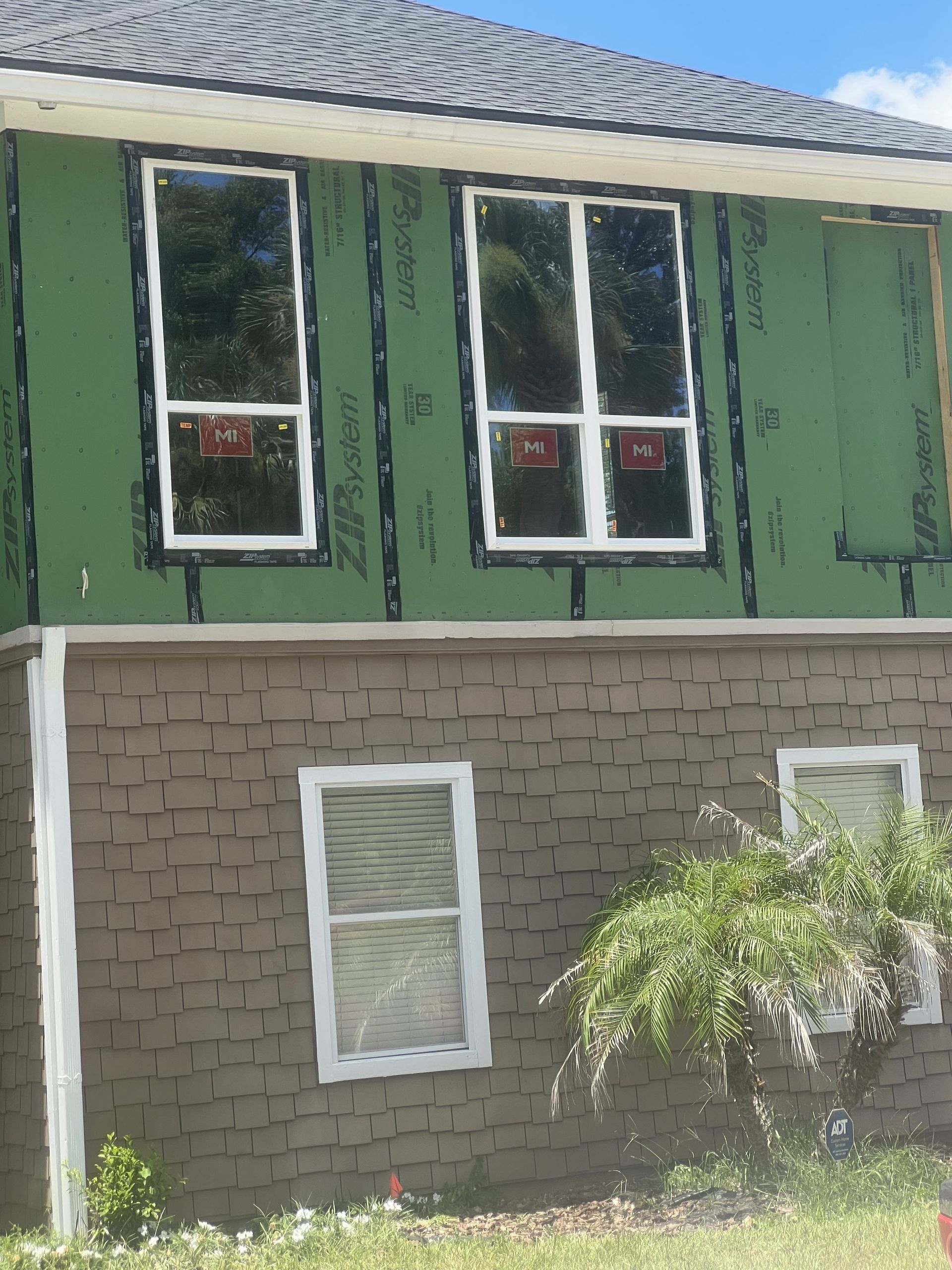 Two-story house with green upper siding, brick lower wall, white-trimmed windows, and a small palm in front
