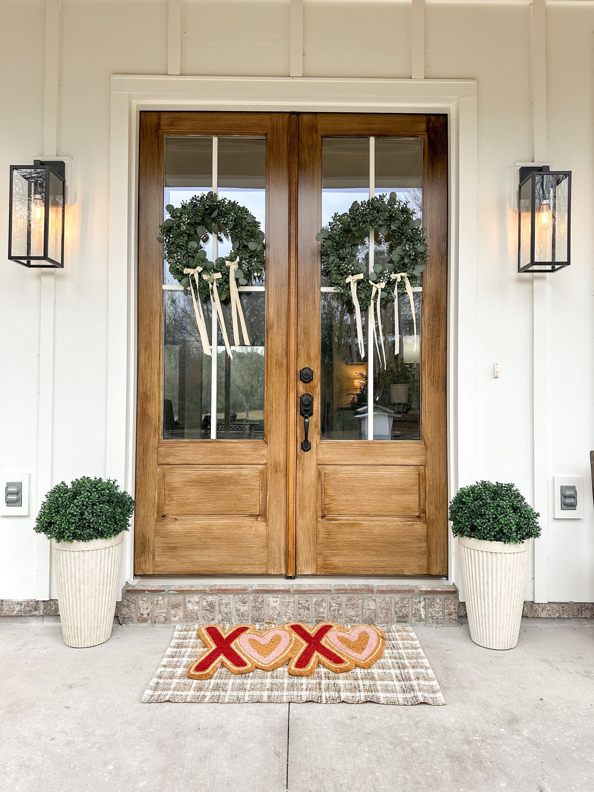 Front porch with wooden double doors, wreaths, lantern sconces, and a red 