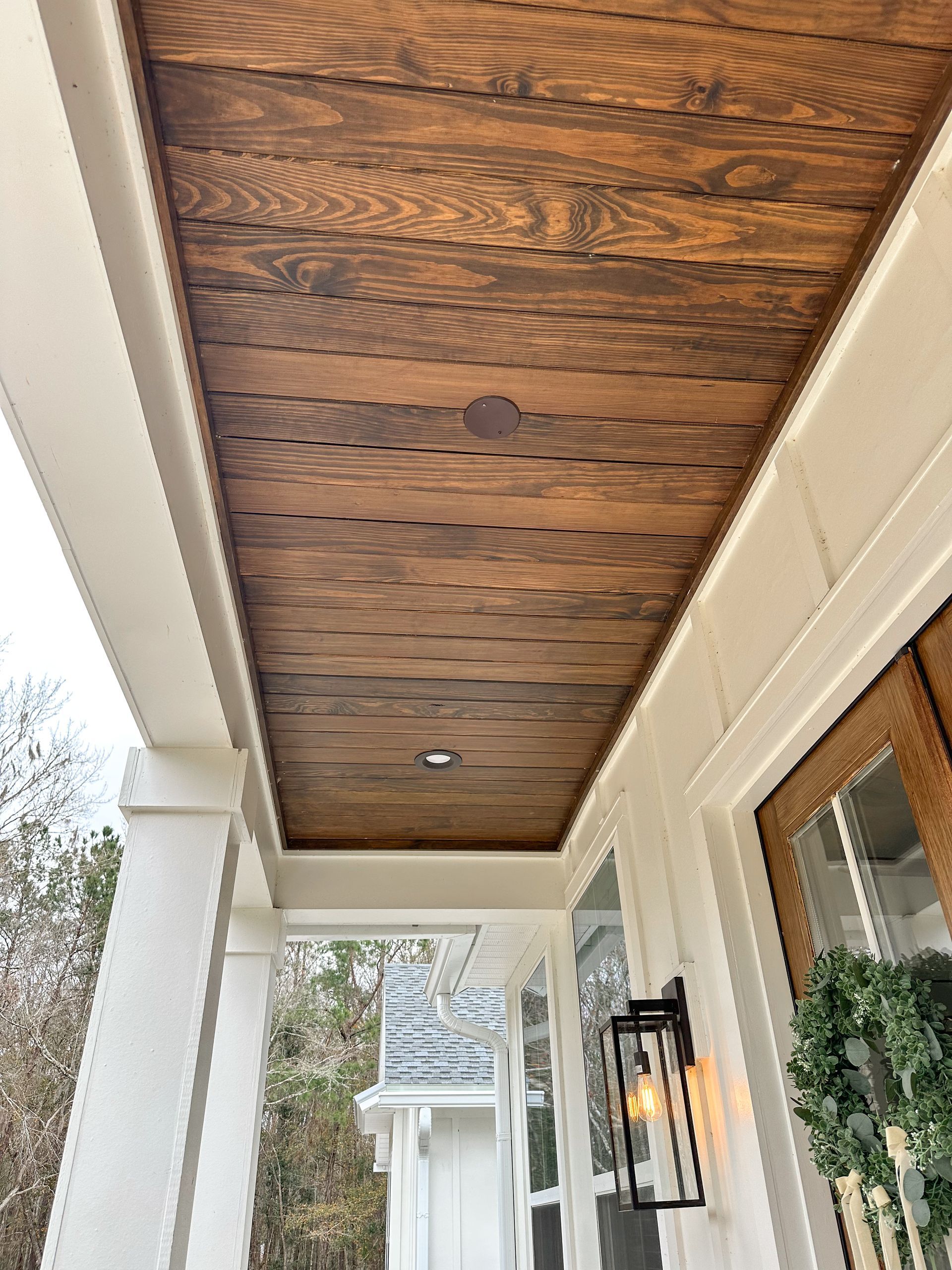 Covered porch with wood-paneled ceiling, white columns, and a wall-mounted lantern beside a glass door.