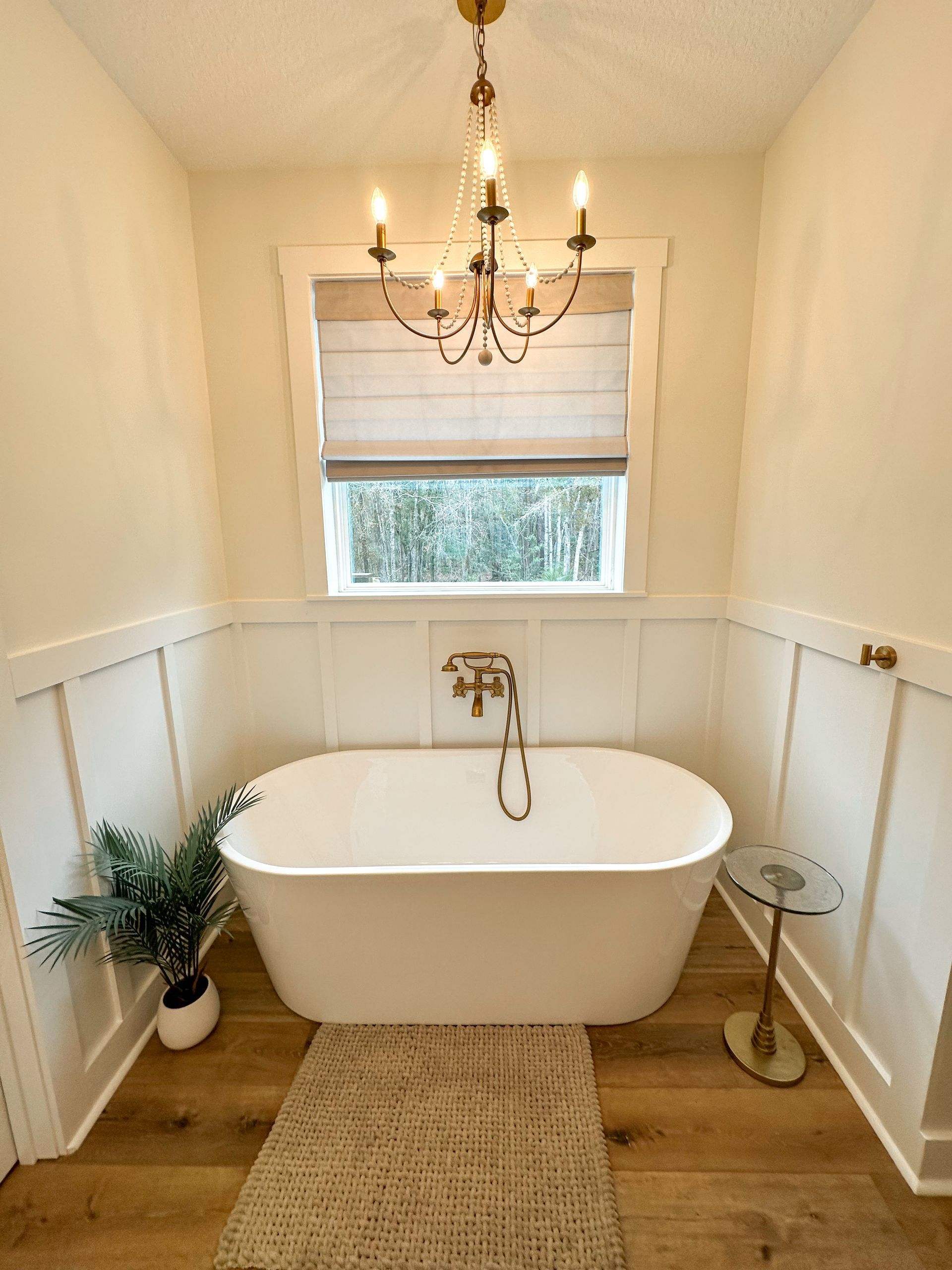 Bright white bathroom with freestanding tub, chandelier, woven rug, and window blind above the tub