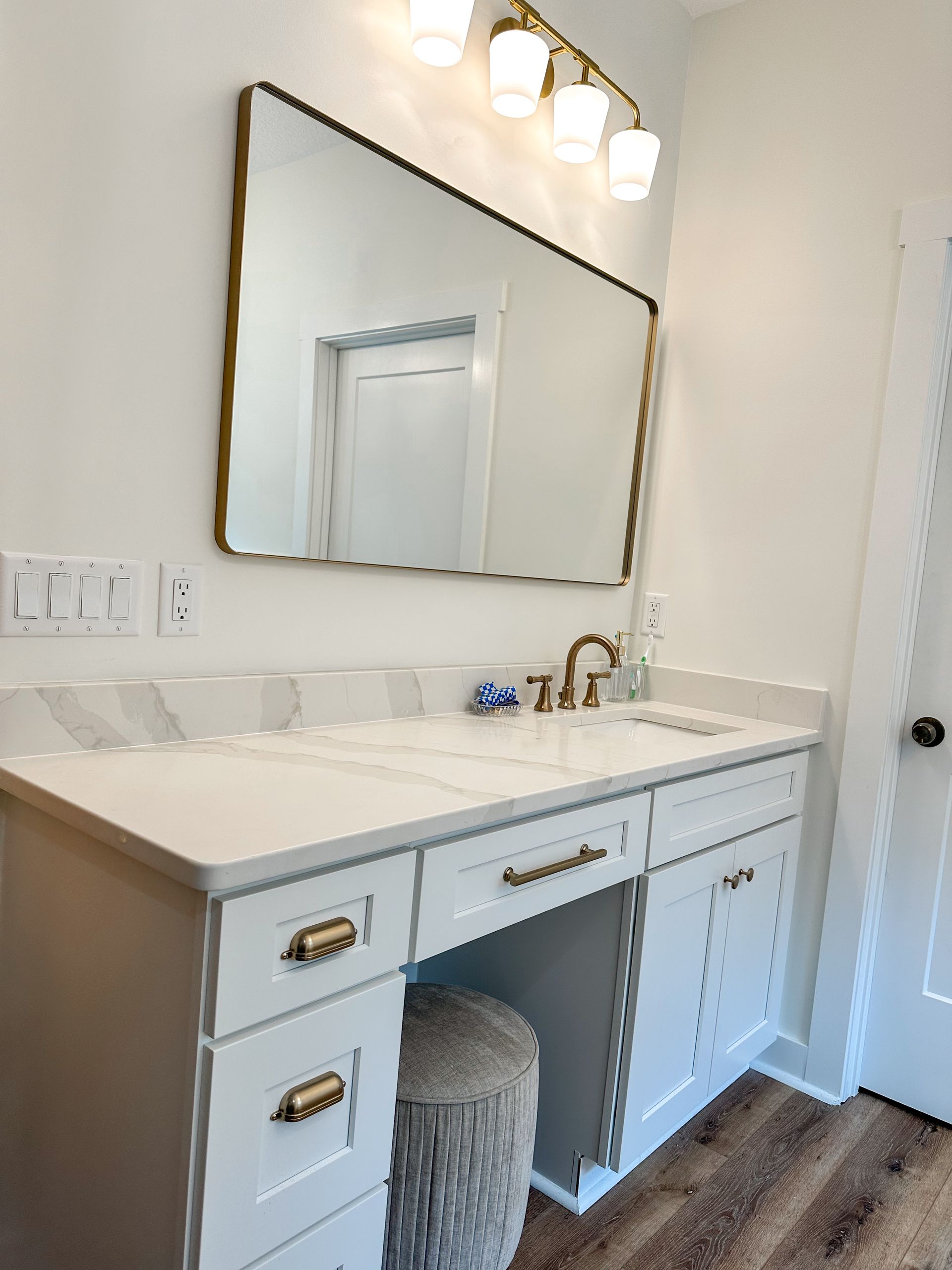 White bathroom vanity with mirror, brass faucet, and wall sconces above a marble-look countertop.