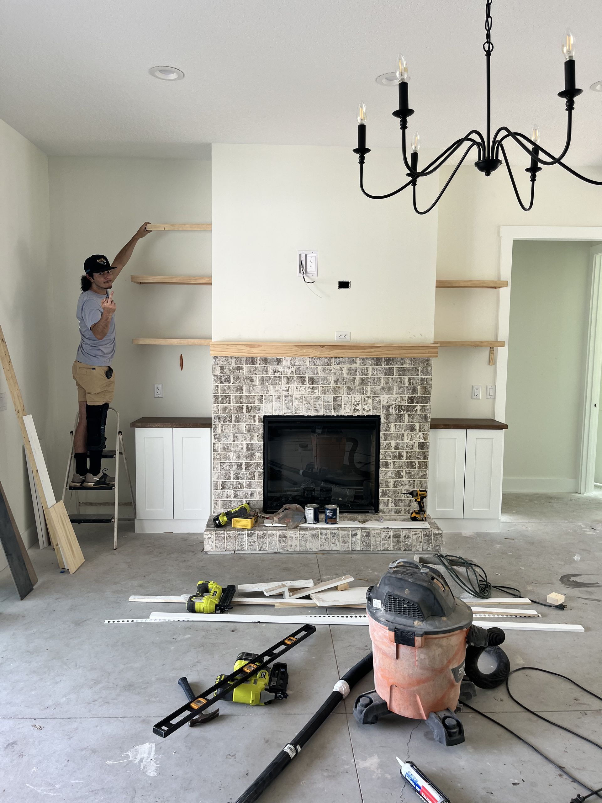 Person on ladder installing shelves in a room with a fireplace and renovation tools scattered on the floor