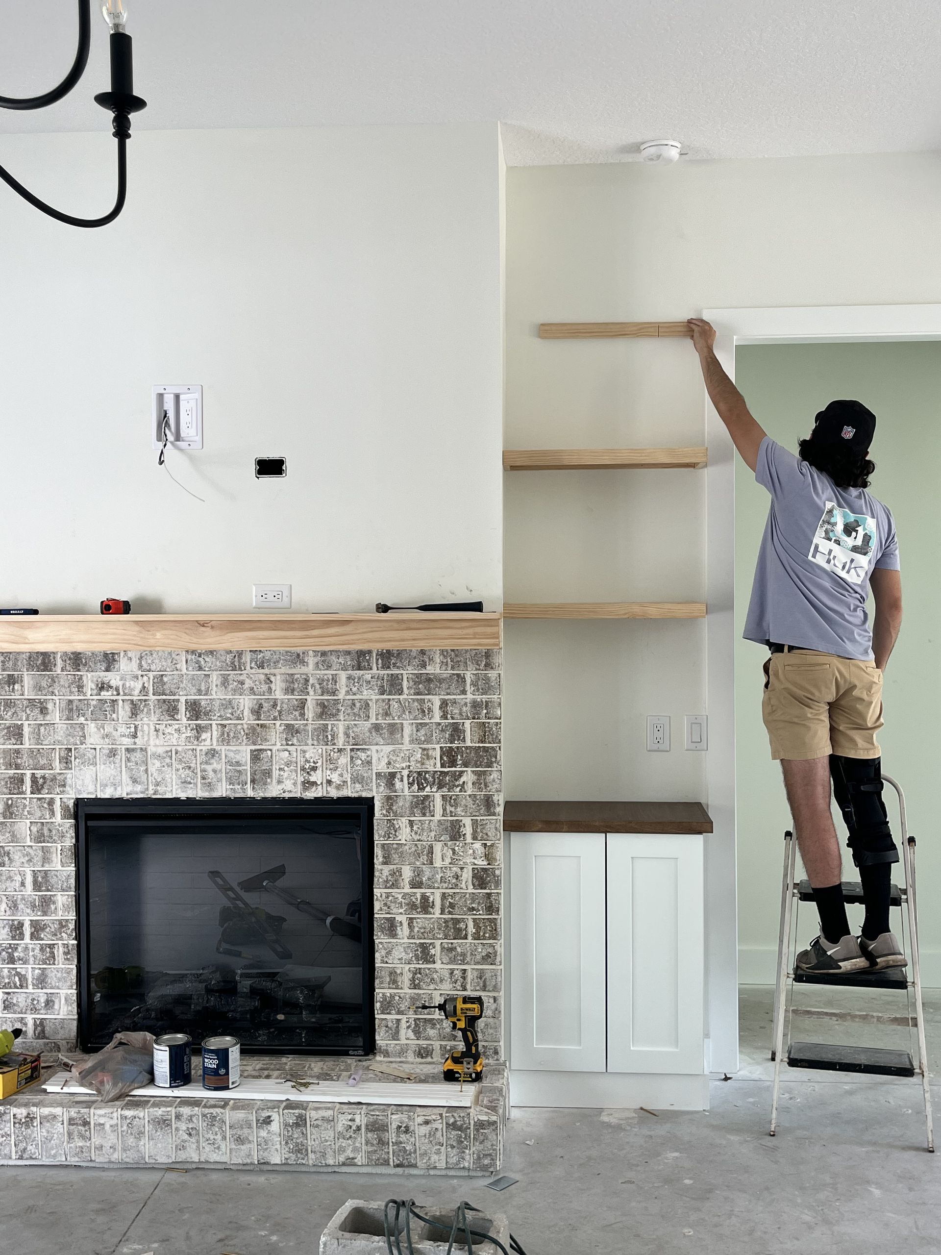 Person on a ladder reaching for shelving beside a fireplace in a white room.