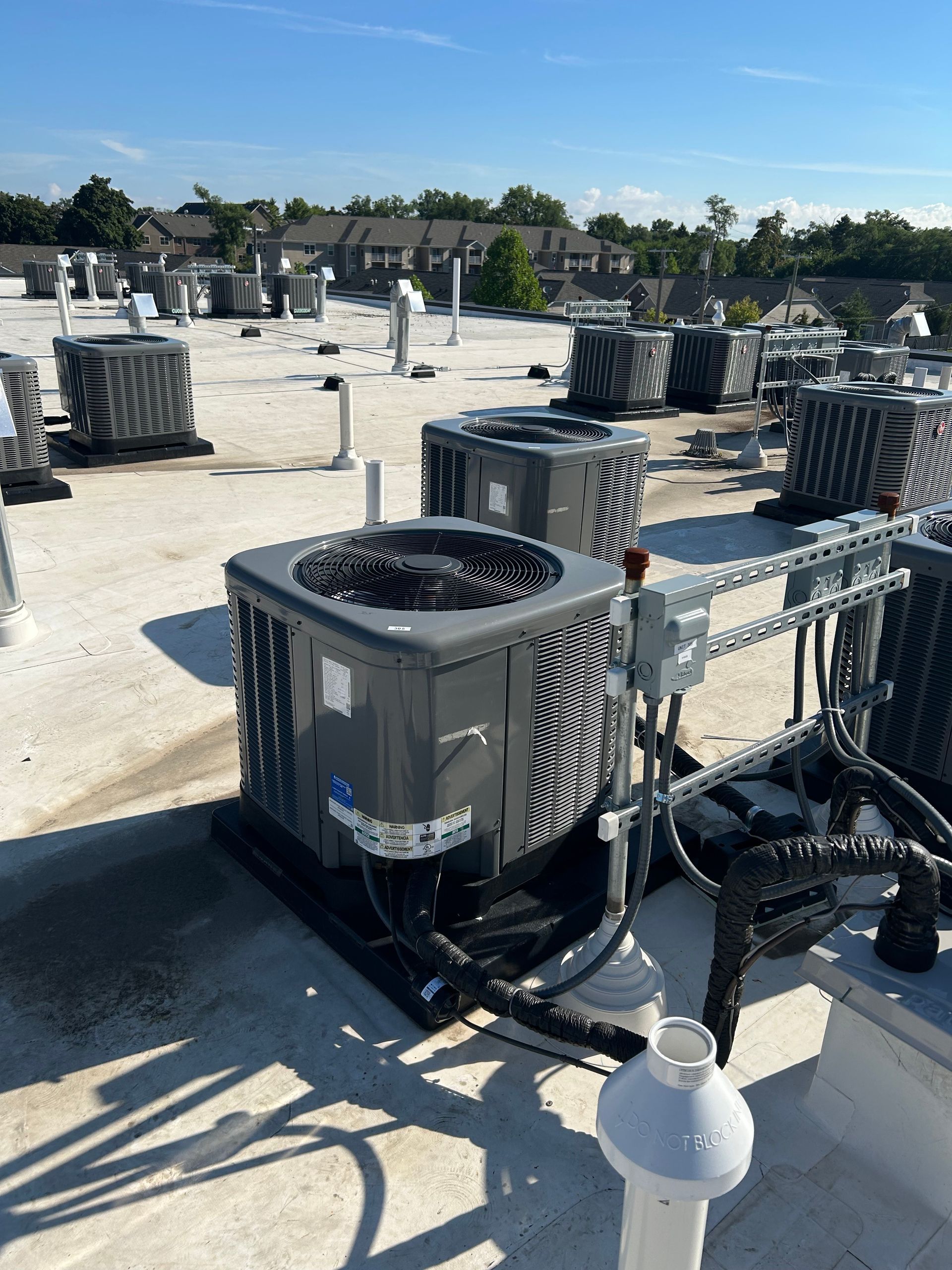 AC units on a rooftop, sunny day. Gray, black units, white roof. Other buildings in background.