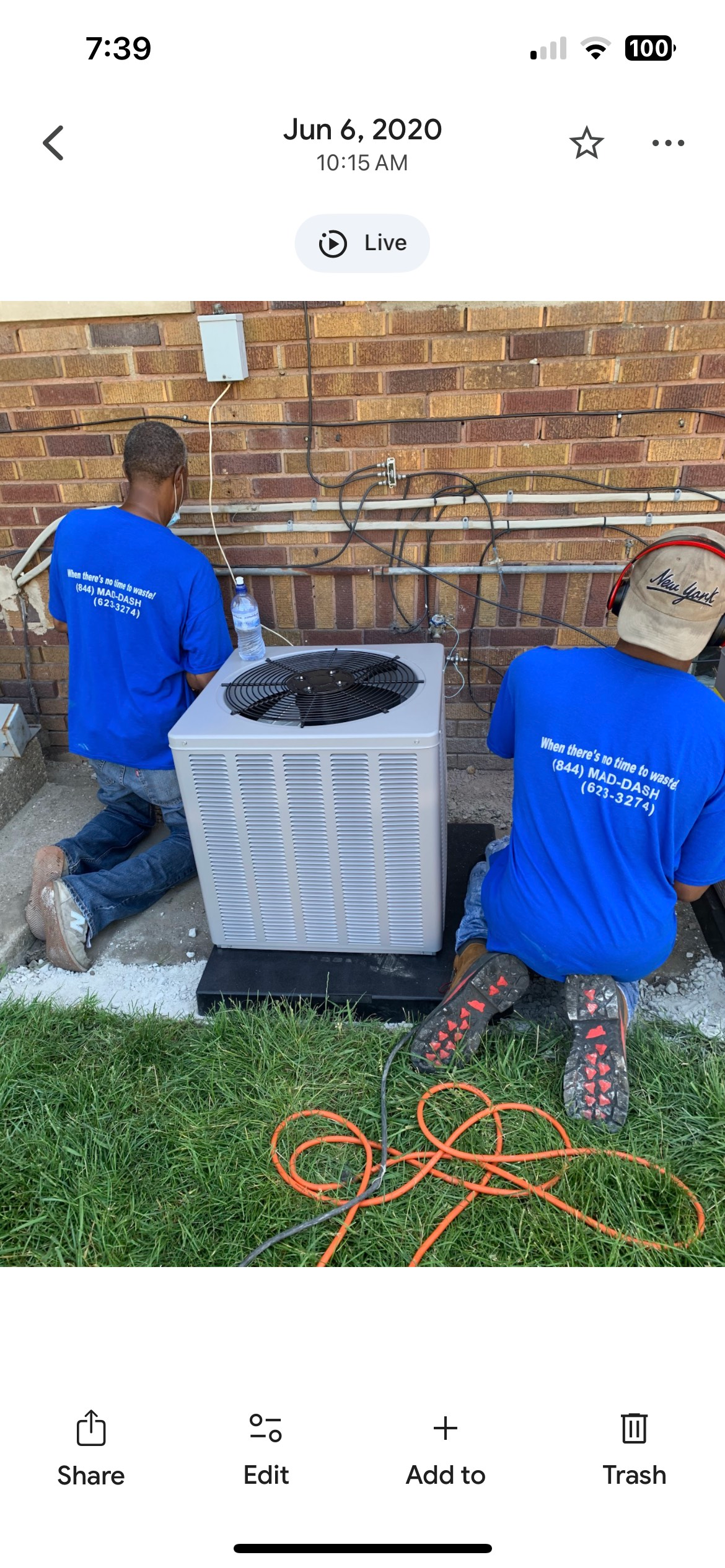 Two workers in blue shirts installing an air conditioning unit outdoors. Brick wall background, green grass below.