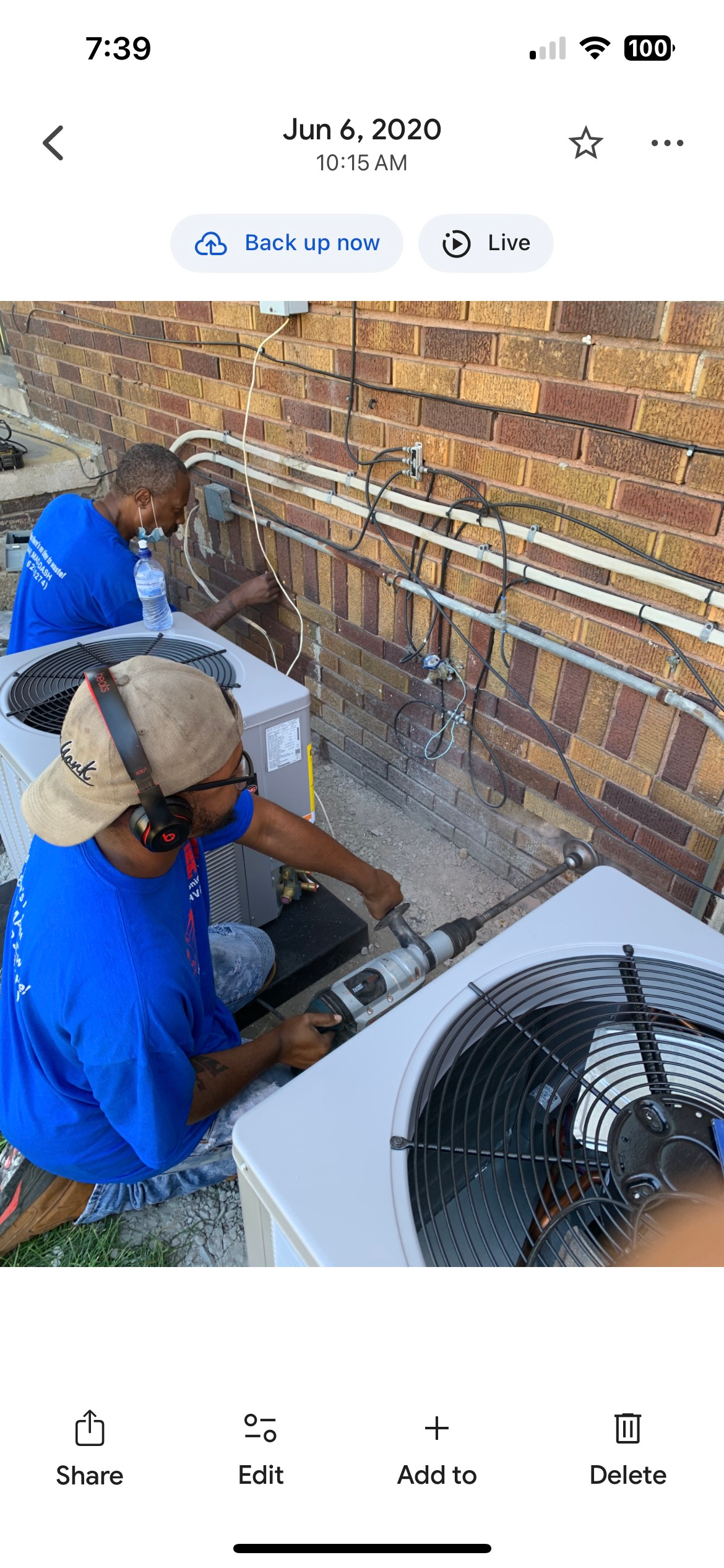 Two workers in blue shirts repair AC units on a rooftop near a brick wall.