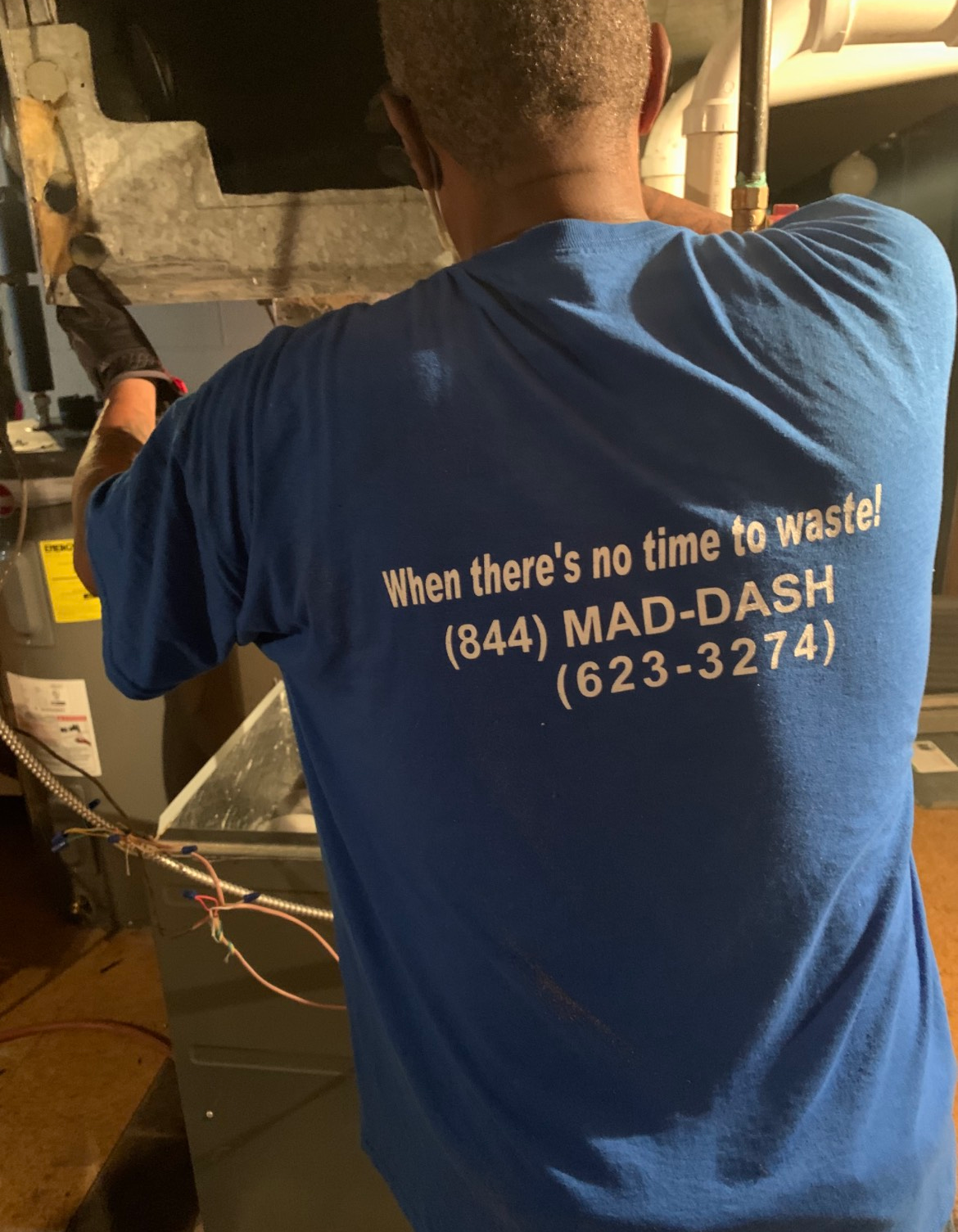 HVAC technician working on furnace, wearing a blue shirt with company logo and phone number.
