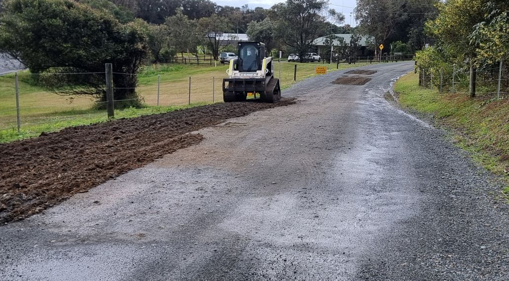 A small tractor levels gravel on a rural road next to a field.