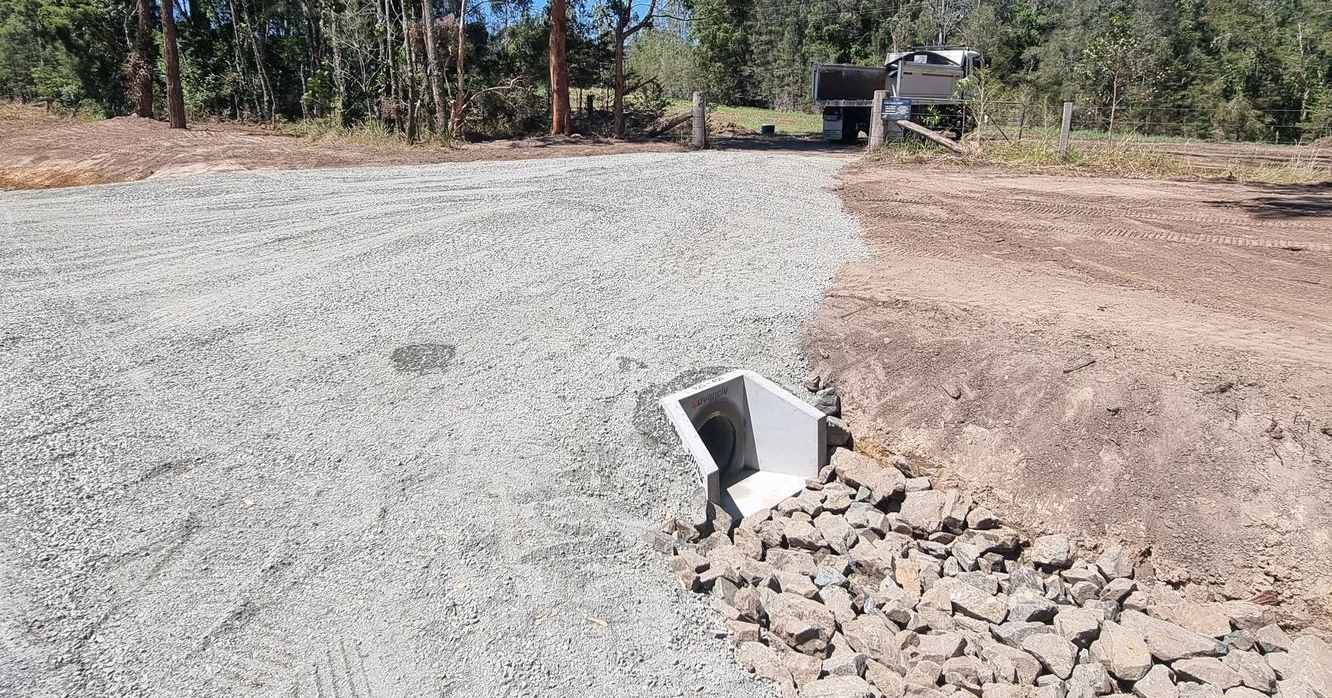 Gravel driveway with a drainage culvert; brown dirt to the right, trees in the background.