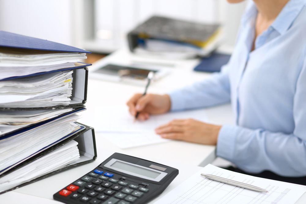 Woman in Blue Shirt Writing at Desk With Calculator and Stacked Binders — Blue Chip Bookkeeping in Cairns, QLD